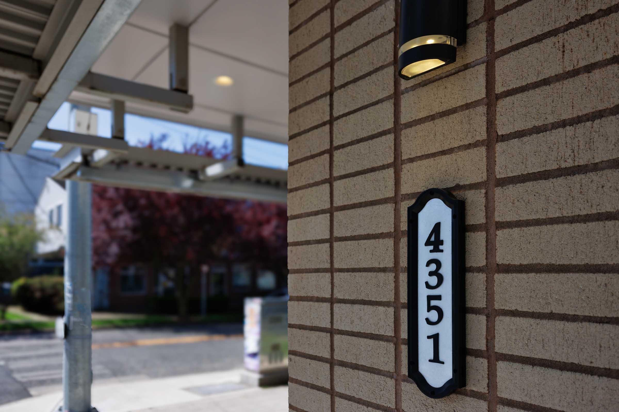 House number 4351 displayed on a brick wall, with a light fixture above it. The background features an awning and blurred greenery, suggesting an outdoor area. The image focuses on the address, with a clear, modern design of the number plate.