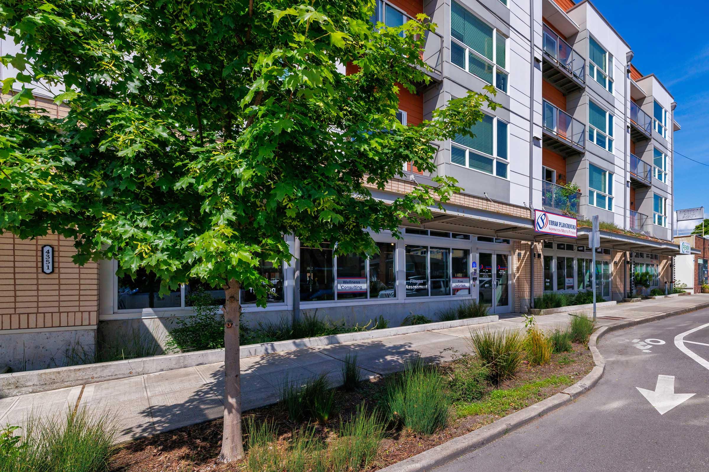 A modern multi-story building with large windows and balconies, surrounded by green landscaping. A tree is visible in the foreground, and there is a sidewalk leading to the building's entrance, which features a sign. The scene is bright and sunny, showcasing a clear blue sky.