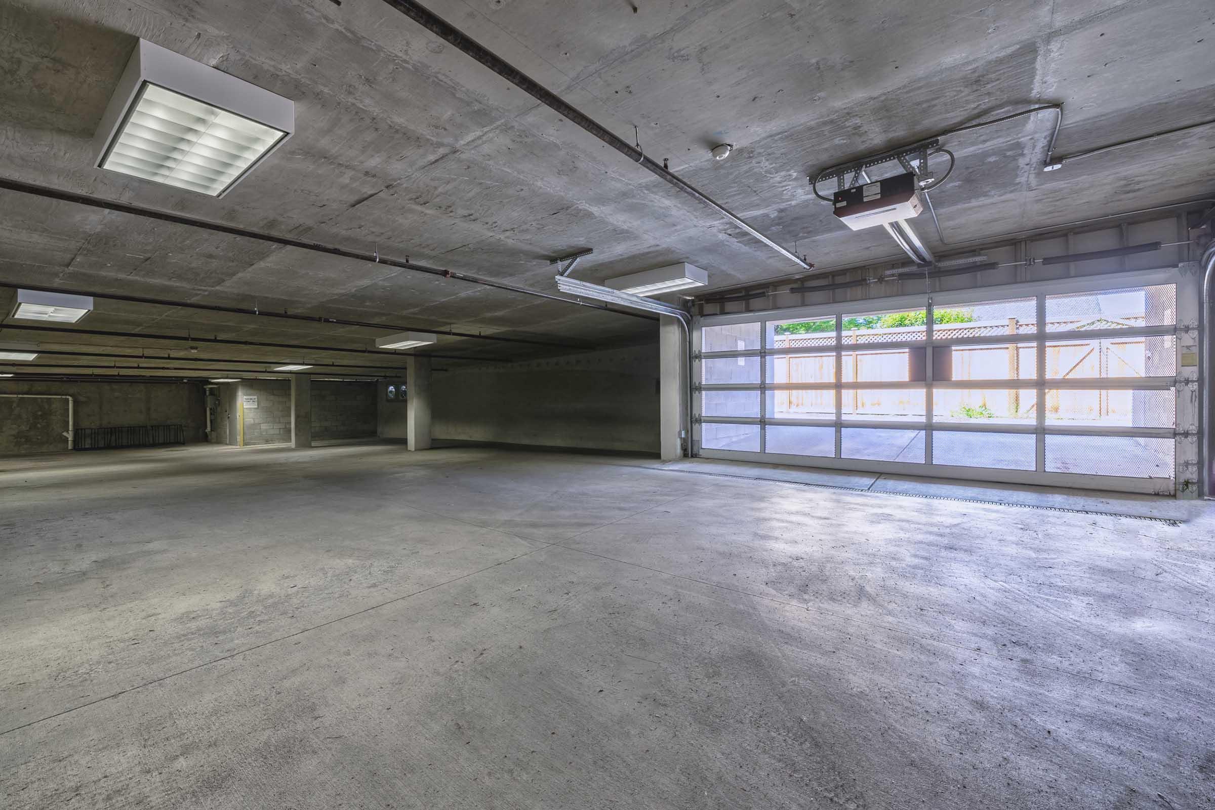 A spacious, empty garage with concrete walls and flooring. Bright fluorescent lights illuminate the area, and a large glass garage door provides access to an outdoor space. The overall atmosphere is clean and modern, with minimal furnishing or equipment present.