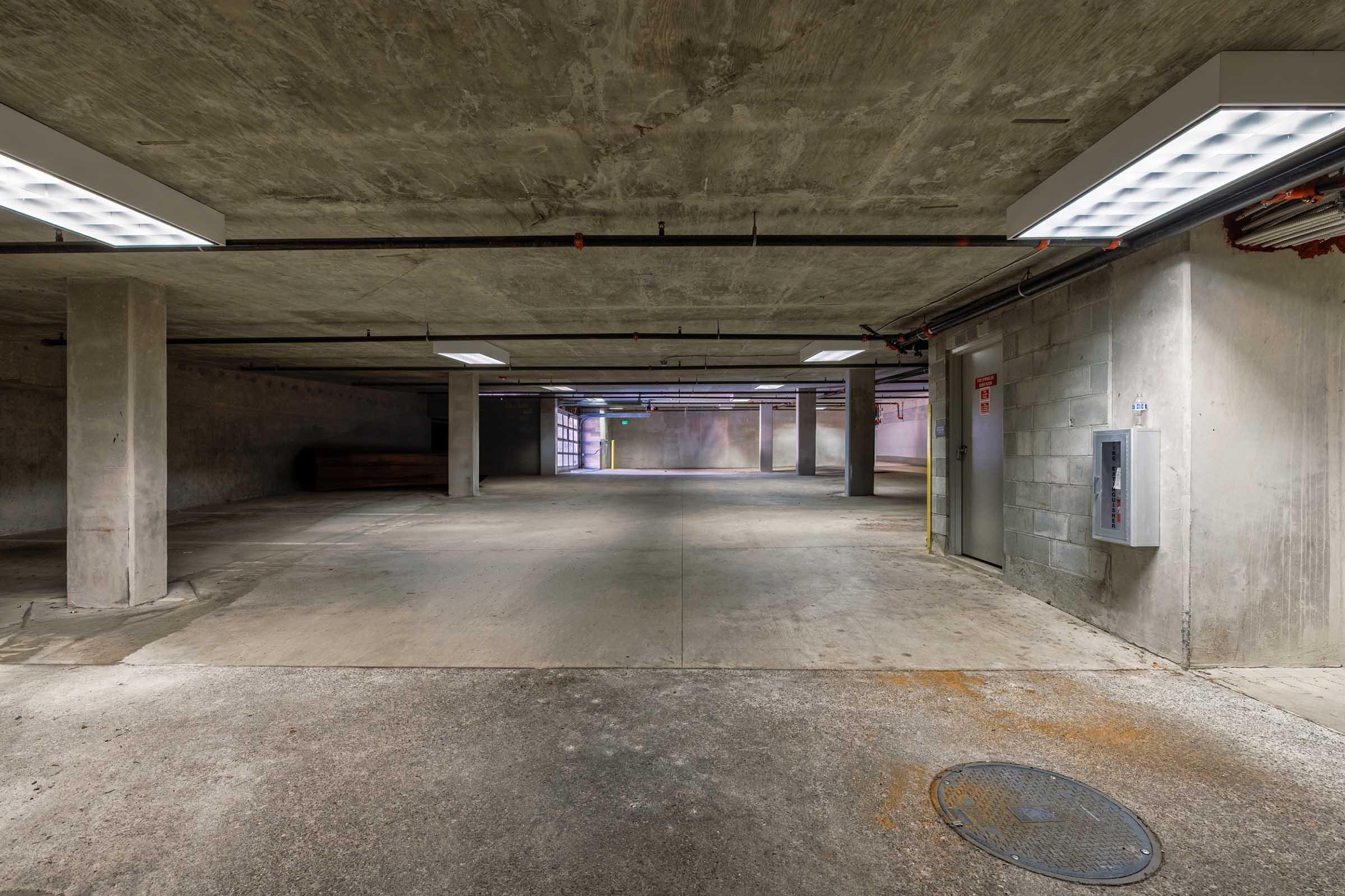 A dimly lit underground parking garage with concrete walls and floors. Several ceiling lights illuminate the space, revealing empty parking spots and exposed beams. An elevator door and a utility box are visible on one wall, along with a drain on the floor. The ambiance is quiet and spacious, with no vehicles present.