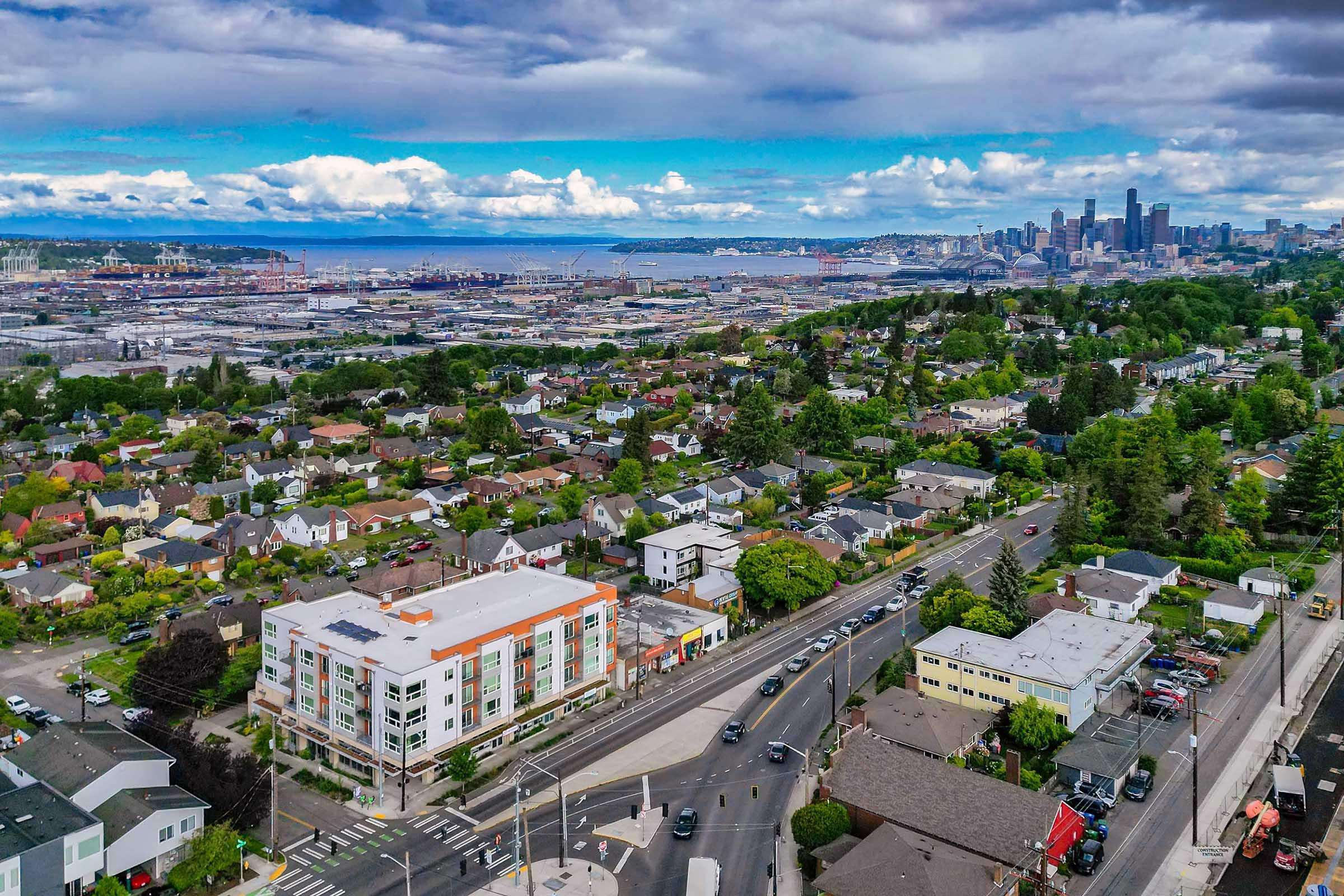 Aerial view of a bustling cityscape featuring residential areas and a commercial building in the foreground, with lush greenery. The background showcases a skyline of tall buildings against a partly cloudy sky, with water and port facilities visible in the distance.