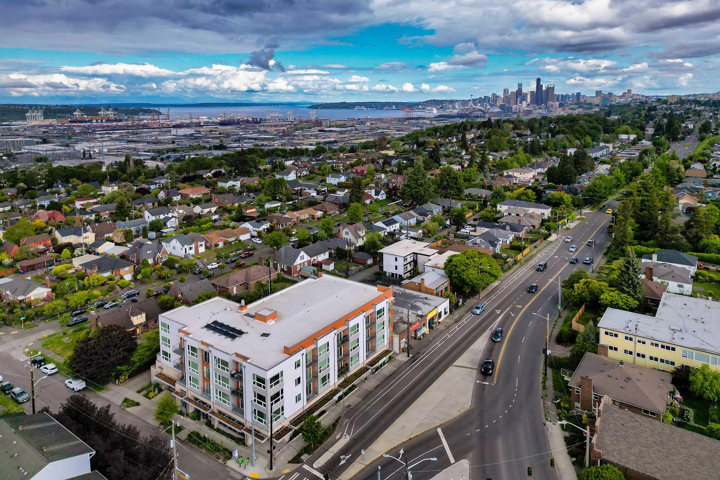 Aerial view of a suburban area with residential homes and a multi-story building in the foreground. A highway runs through the scene, leading towards a city skyline in the background, featuring tall buildings against a cloudy sky. The landscape shows greenery and urban infrastructure.