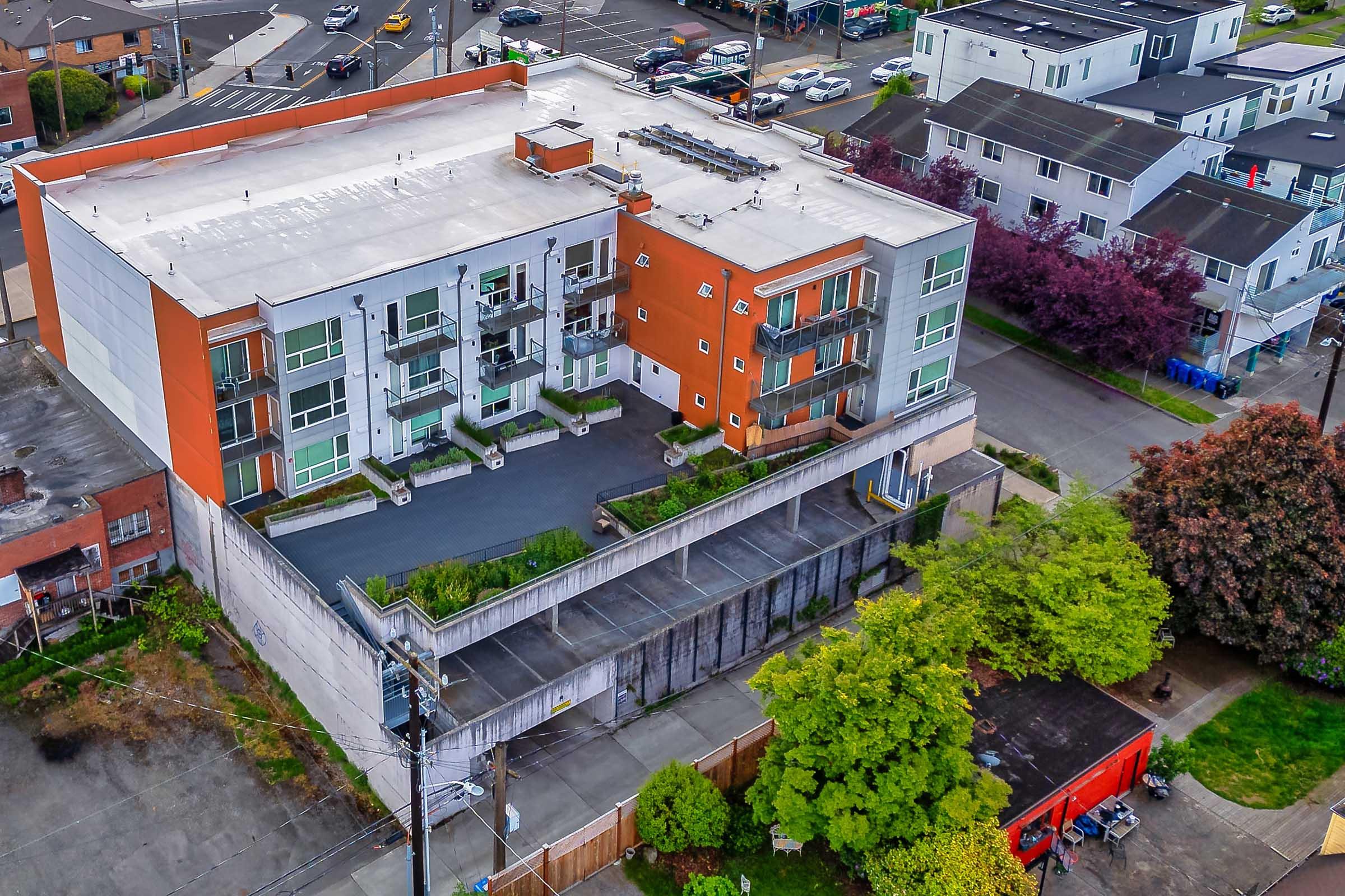 Aerial view of a modern multi-unit residential building featuring a mix of orange and white façades. Surrounding the structure are landscaped areas and parking spaces, with nearby streets, trees, and other buildings visible in the background. The setting appears urban and well-maintained.
