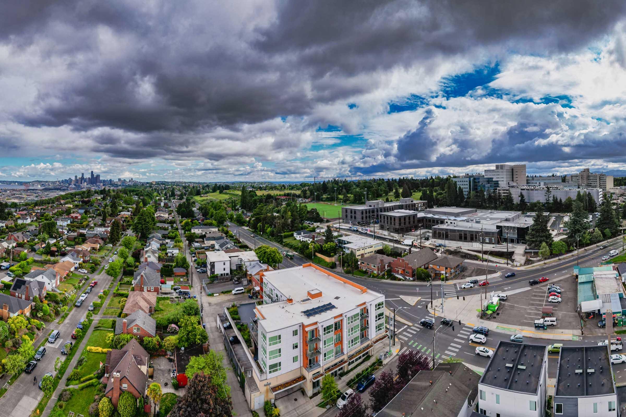 Aerial view of a suburban area featuring a mix of residential homes, commercial buildings, and green parks. The skyline of a city is visible in the distance under a dramatic sky filled with clouds. Roads and cars are seen winding through the neighborhood, highlighting urban development amidst nature.