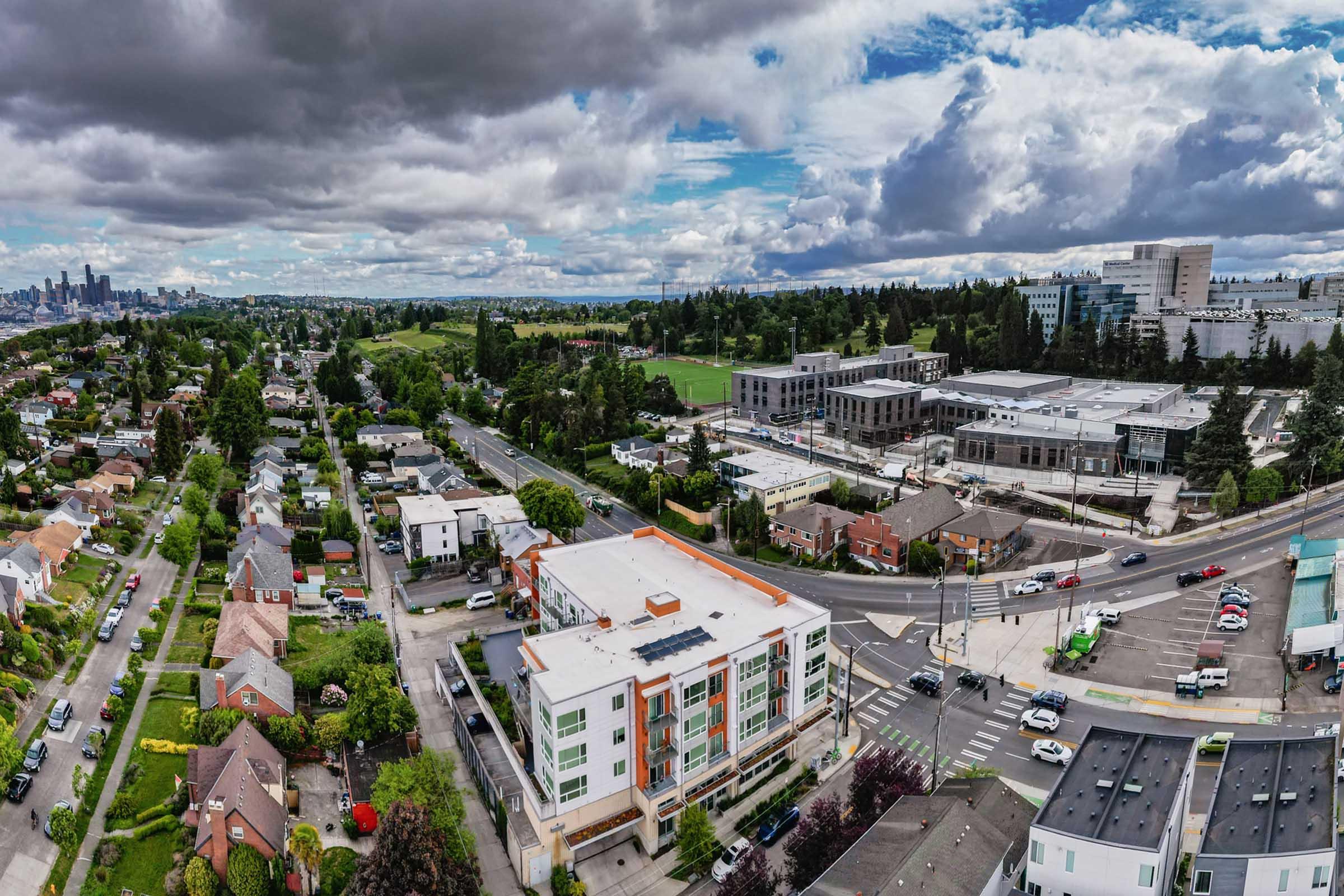 Aerial view of a suburban area featuring residential buildings and modern commercial structures. Lush green spaces and parks are visible, along with scattered cars and roads. In the distance, a city skyline is partially visible under a cloudy sky, suggesting a vibrant urban setting combined with nature.