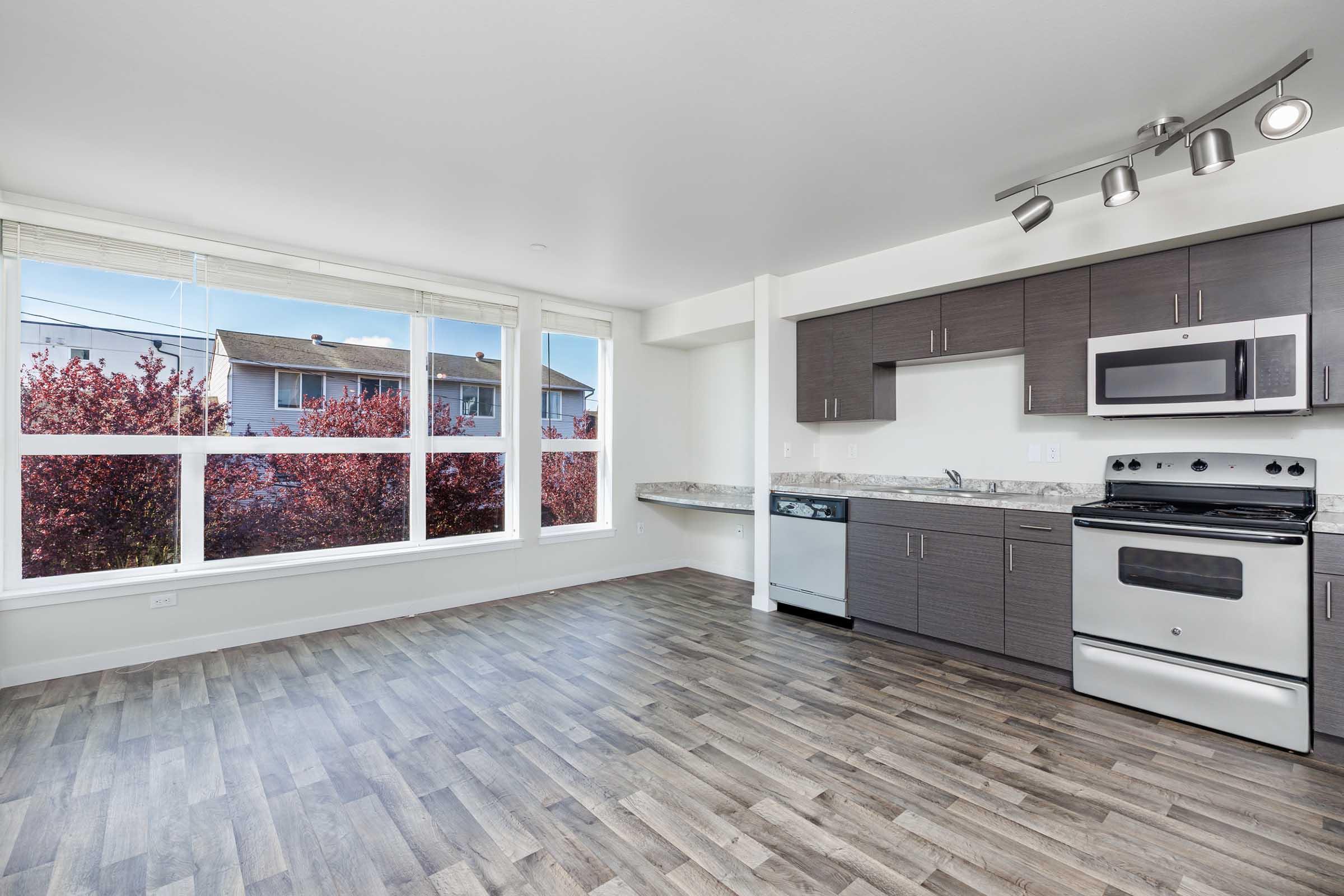 A modern kitchen and living area featuring sleek gray cabinetry, stainless steel appliances, and large windows with a view of red-leaved trees outside. The space has a light and airy atmosphere with wood-like flooring and ample natural light.