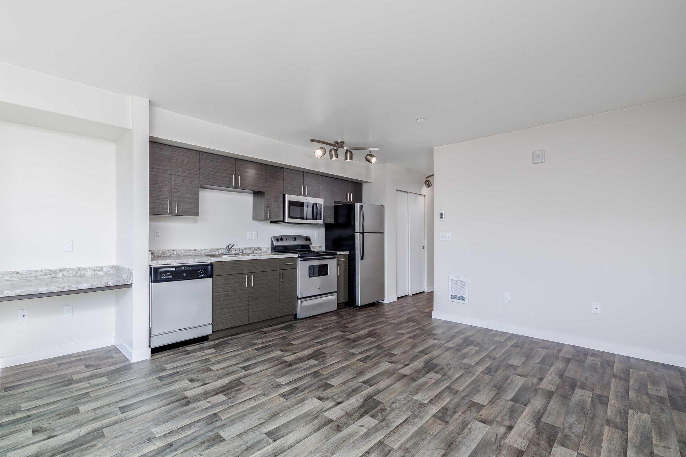 Modern kitchen featuring stainless steel appliances, including a refrigerator, oven, and dishwasher. The design includes dark wood cabinetry and a granite countertop, with a spacious open layout and laminate flooring that extends into the living area. Natural light brightens the space.