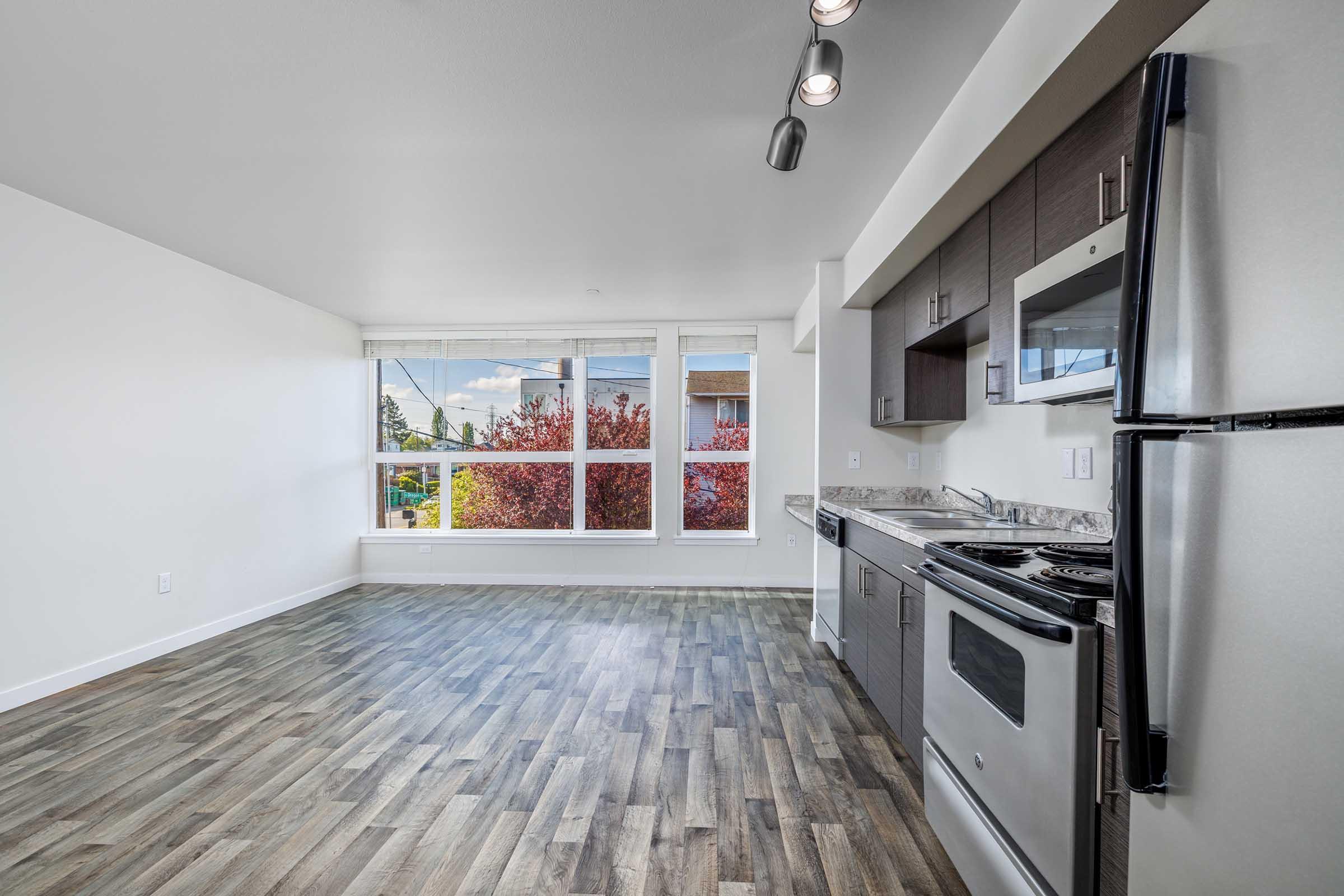 Spacious kitchen and living area with modern appliances, including a stove and refrigerator. Large windows allow natural light, showing a view of vibrant red trees outside. The flooring is trendy and wooden, enhancing the contemporary feel of the space.