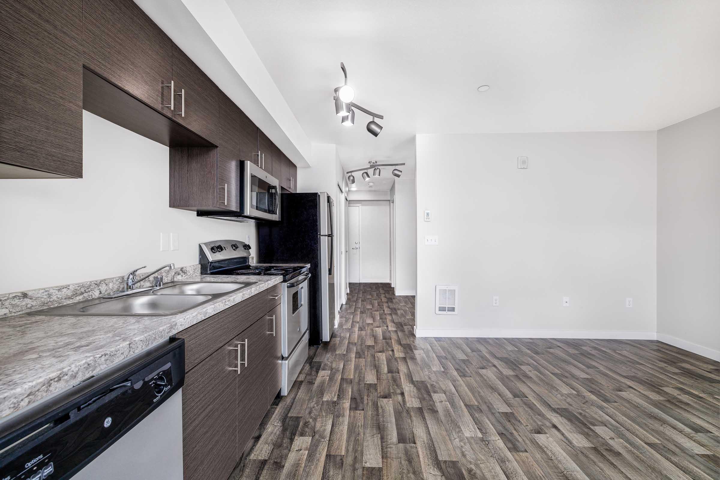A modern kitchen layout featuring stainless steel appliances, including a stove, refrigerator, and dishwasher, set against dark cabinetry. The space has a granite countertop and sleek cabinetry, with a visible hallway leading to another room and light-colored walls enhancing the bright atmosphere.