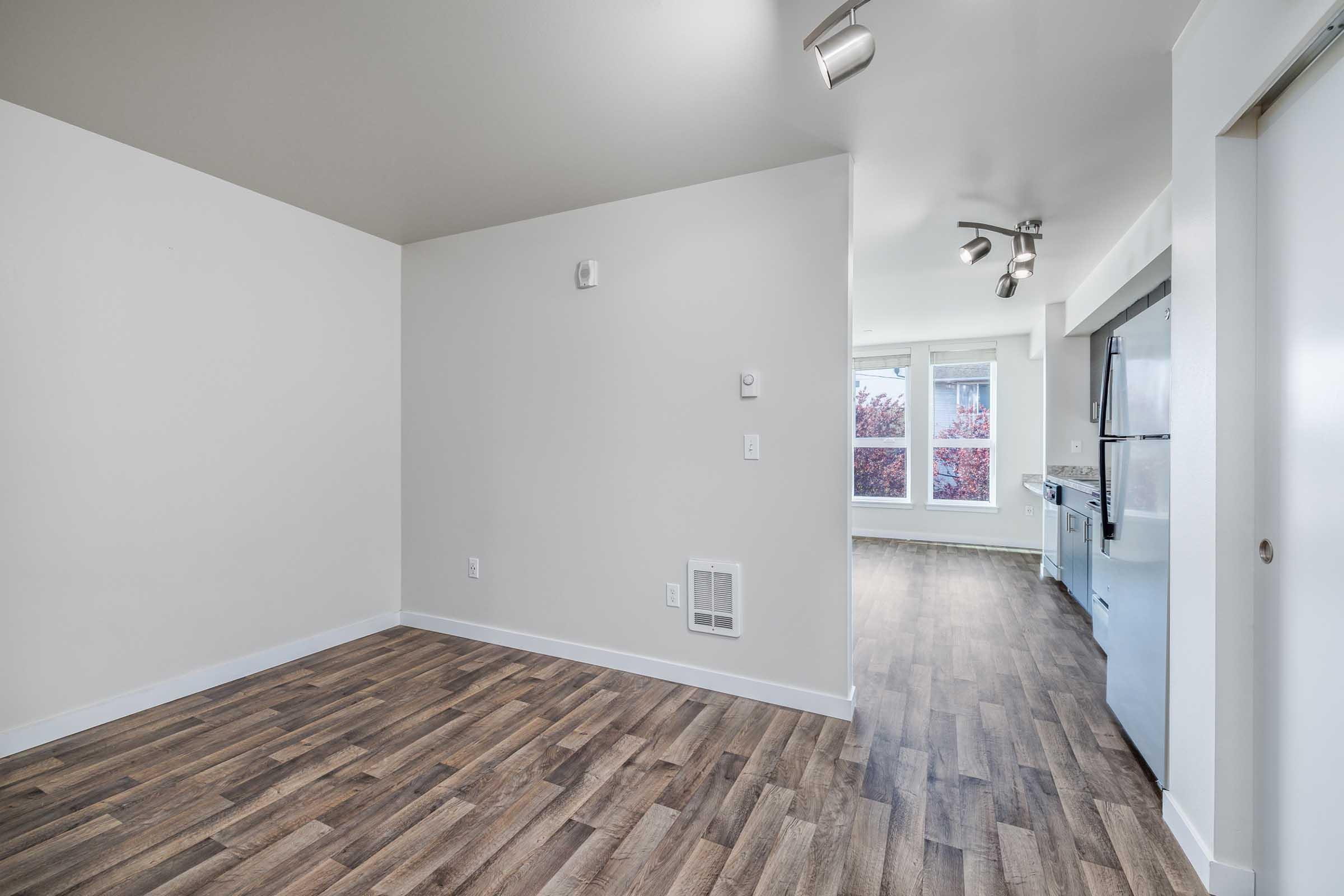 A spacious, modern apartment interior featuring light-colored walls, laminate flooring, and windows allowing natural light. The room is partially divided, with a kitchen area visible in the background and track lighting installed in the ceiling.