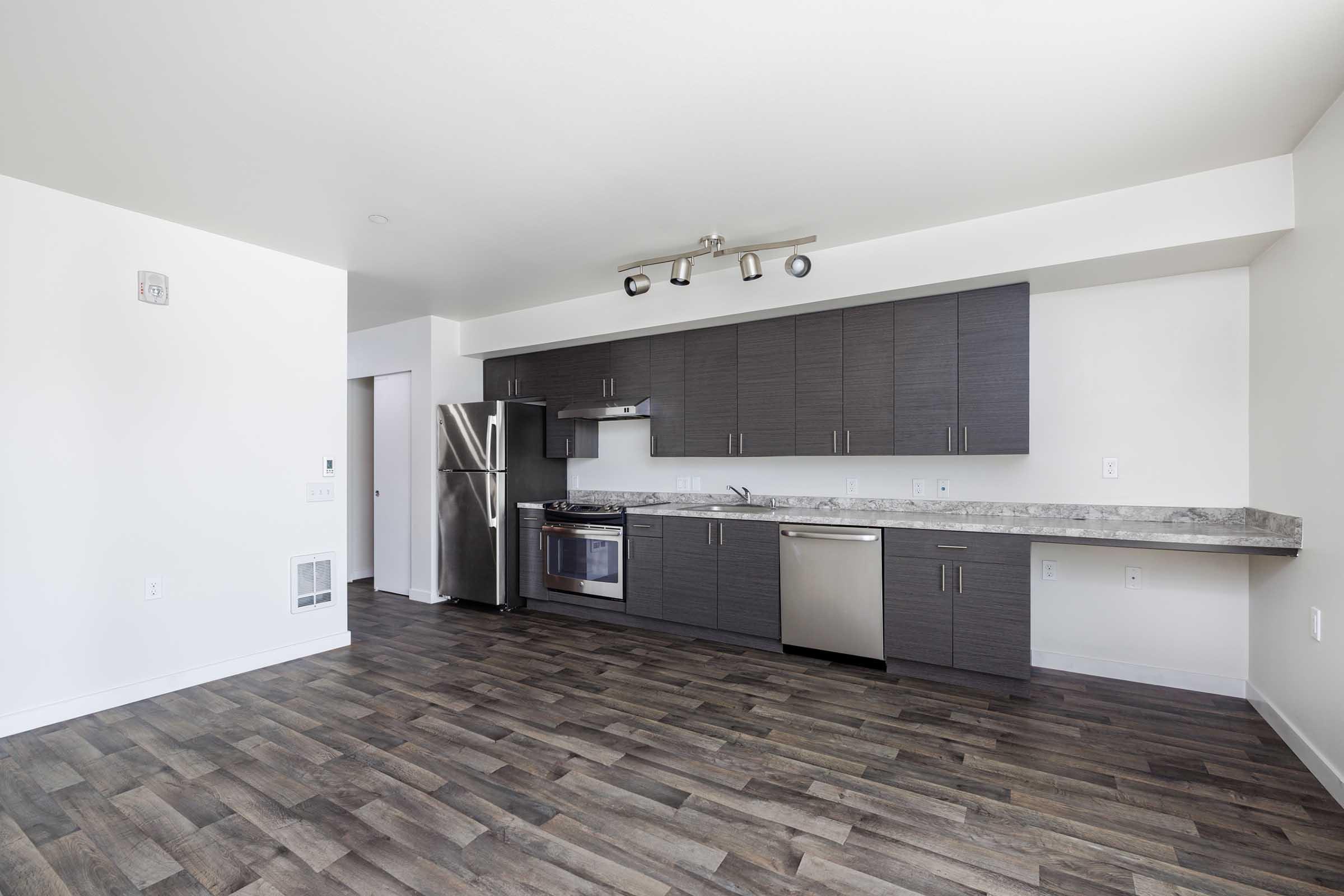 Modern kitchen with dark wood cabinets, stainless steel appliances including a refrigerator and oven, and a stone countertop. It features a spacious layout with a neutral color palette and wood-style flooring, allowing for plenty of natural light.