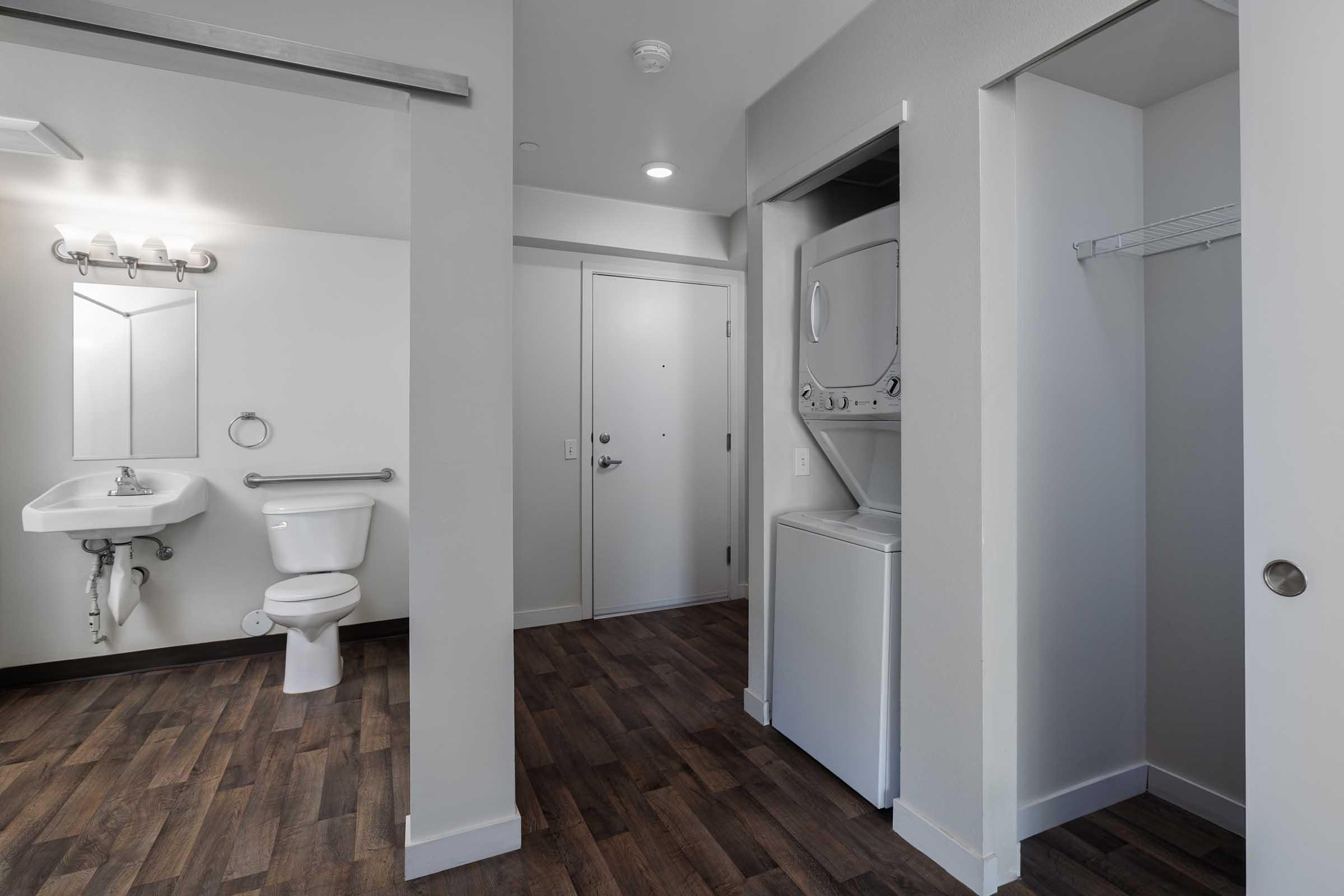 A modern bathroom area featuring a toilet, a small sink with a mirror above, and a stackable washer and dryer unit. The walls are light-colored and the flooring is wood-like. There is a door leading outside and a closet space visible in the corner.