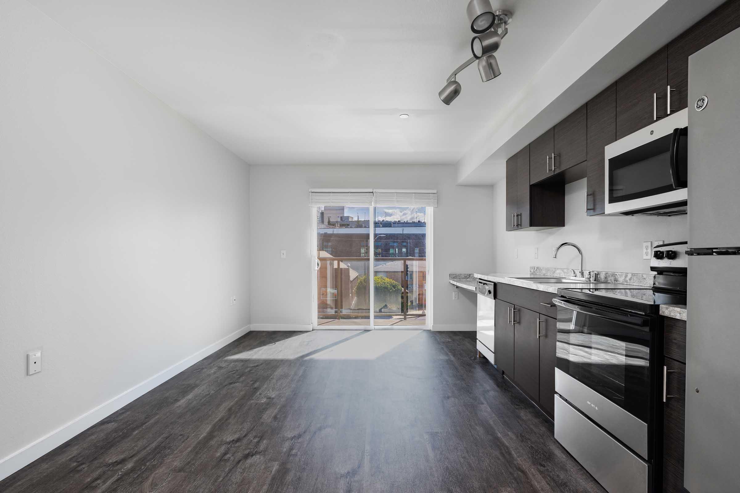 A modern kitchen in an apartment featuring dark cabinetry, a stainless steel refrigerator and microwave, a black oven, and a marble countertop. There is a large window with natural light illuminating the space and dark laminate flooring throughout. The walls are painted white, creating a bright and open atmosphere.