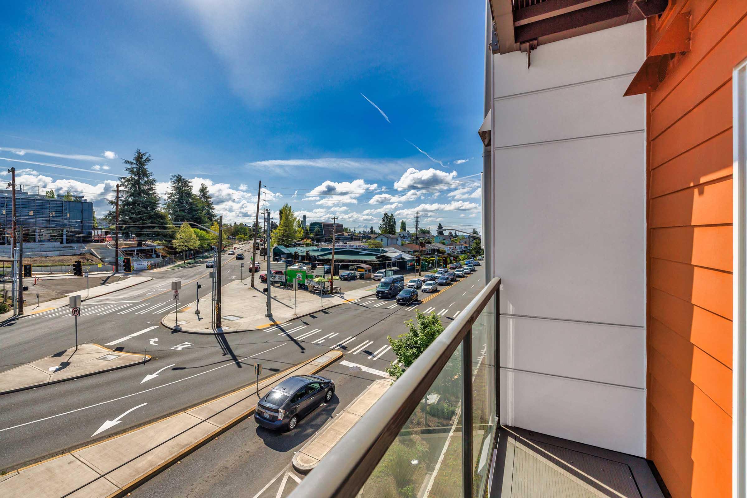 View from a balcony overlooking a busy street intersection. There are several vehicles on the road, trees lining the streets, and a bright blue sky with some clouds. The building features an orange wall on the right side, adding a modern touch to the scene.
