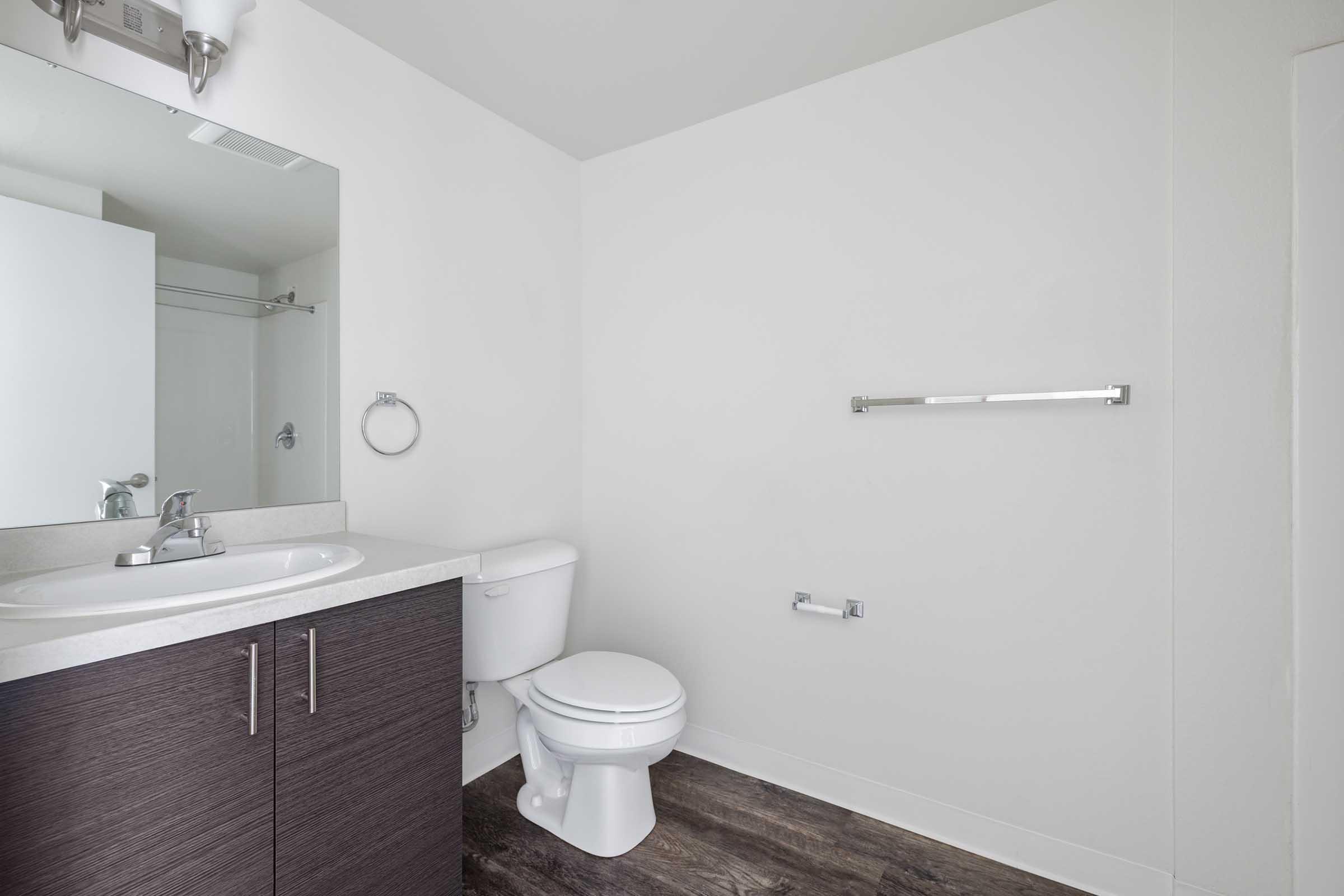 A clean, modern bathroom featuring a white toilet, a simple sink with a faucet, a mirror above the sink, and a towel bar on the wall. The cabinetry is dark wood, and the flooring is a light brown wood texture. The walls are painted white, creating a bright and spacious atmosphere.