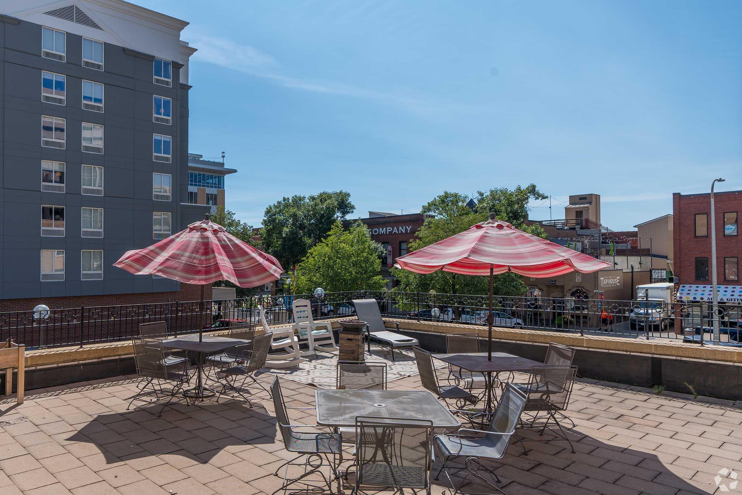 Outdoor seating area with metal tables and chairs, shaded by red and white striped umbrellas. In the background, there's a view of a cityscape featuring buildings and trees under a clear blue sky. The setting appears inviting and suitable for relaxation or social gatherings.