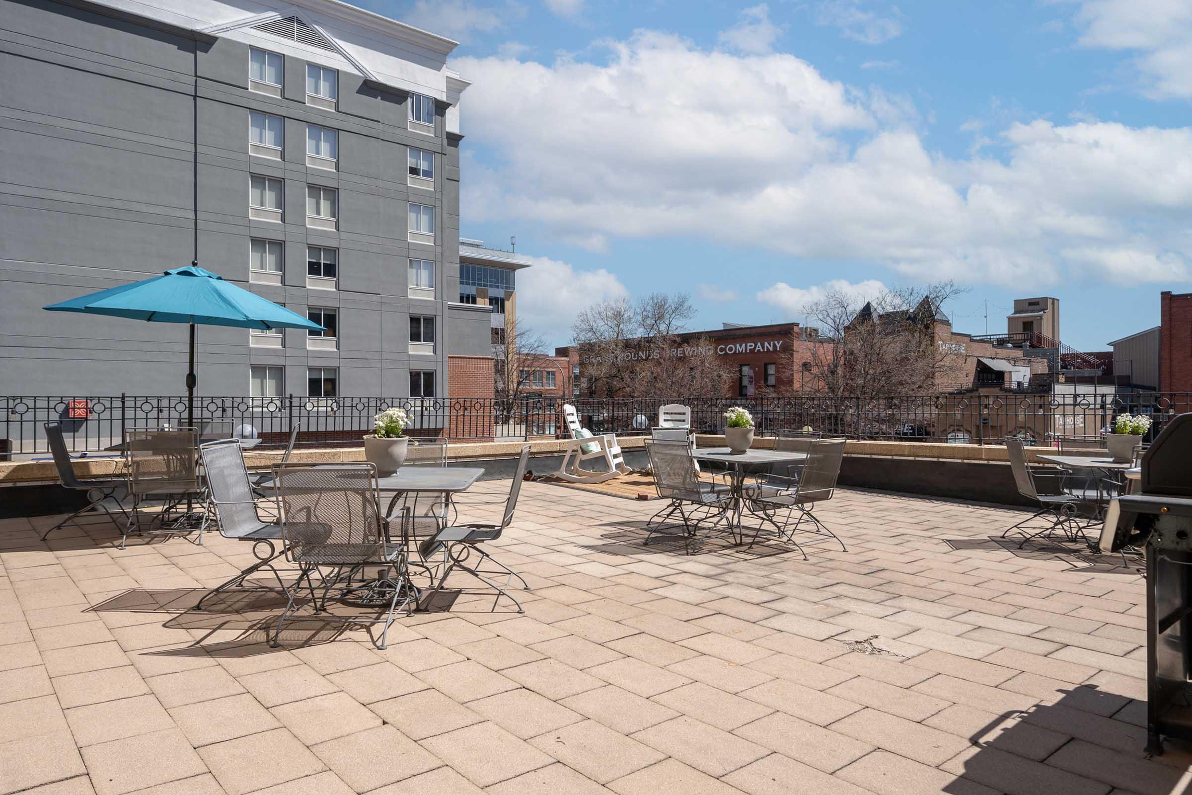 A rooftop terrace featuring several metal tables and chairs, a light blue umbrella, and potted plants. In the background, a gray building and a red brick structure can be seen under a partly cloudy sky. The terrace is paved with beige tiles, creating a spacious and inviting outdoor area.