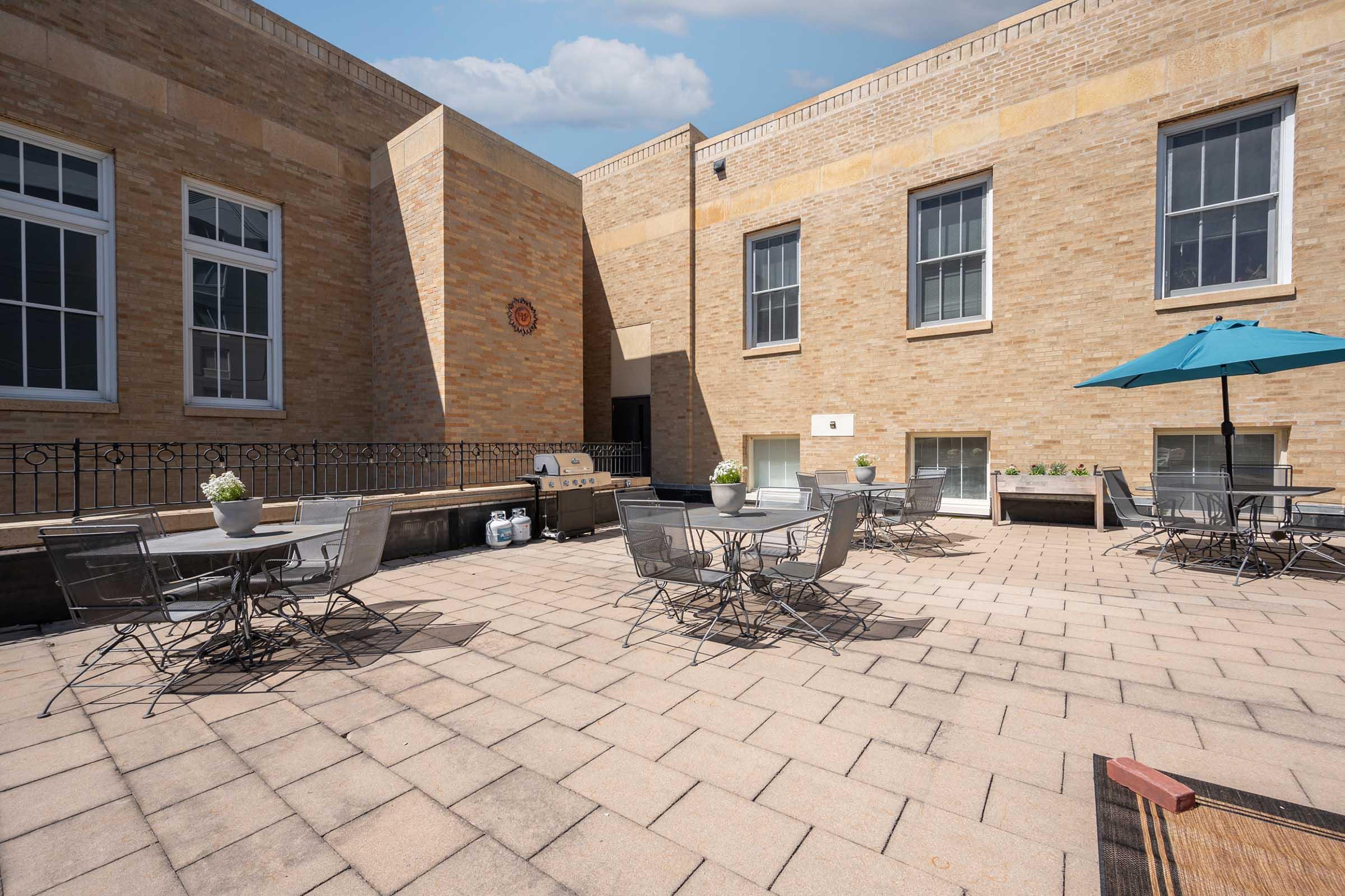 An outdoor patio area featuring several metal tables and chairs, brick walls, and an umbrella providing shade. There is a barbecue grill nearby, along with potted plants on the tables, creating a relaxed atmosphere for gatherings or outdoor dining.