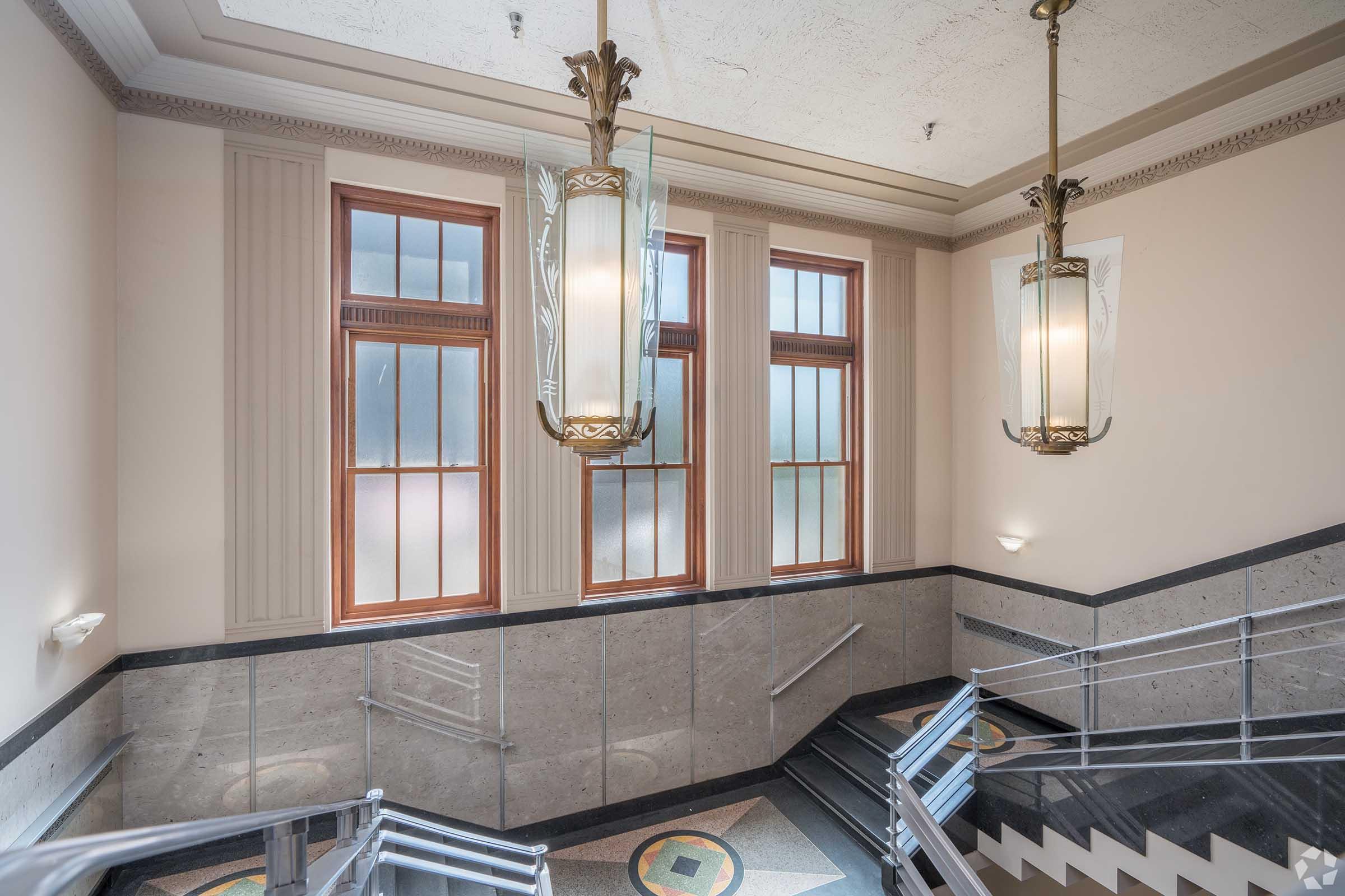 A well-lit stairwell featuring large windows, elegant light fixtures, and decorative molding. The walls are adorned with a light-colored material, and the staircase has a sleek design with metal railings. The overall ambiance is classic and sophisticated, highlighting architectural details.