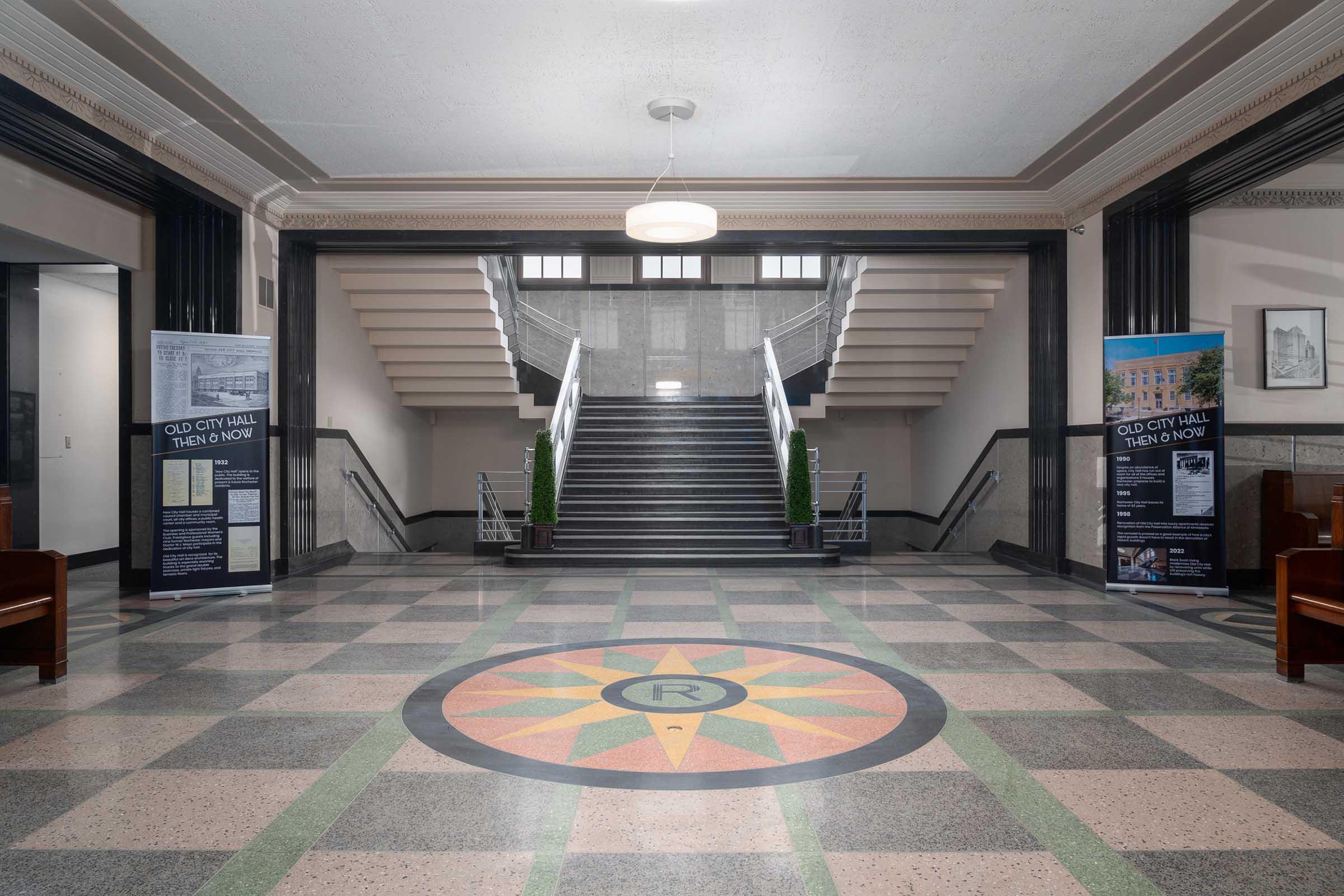Interior view of a historic building featuring a spacious foyer with a large decorative floor compass design. There are two banners on either side, and a grand staircase leads to the upper level, flanked by plants. The walls are adorned with classic architectural details, creating an elegant atmosphere.