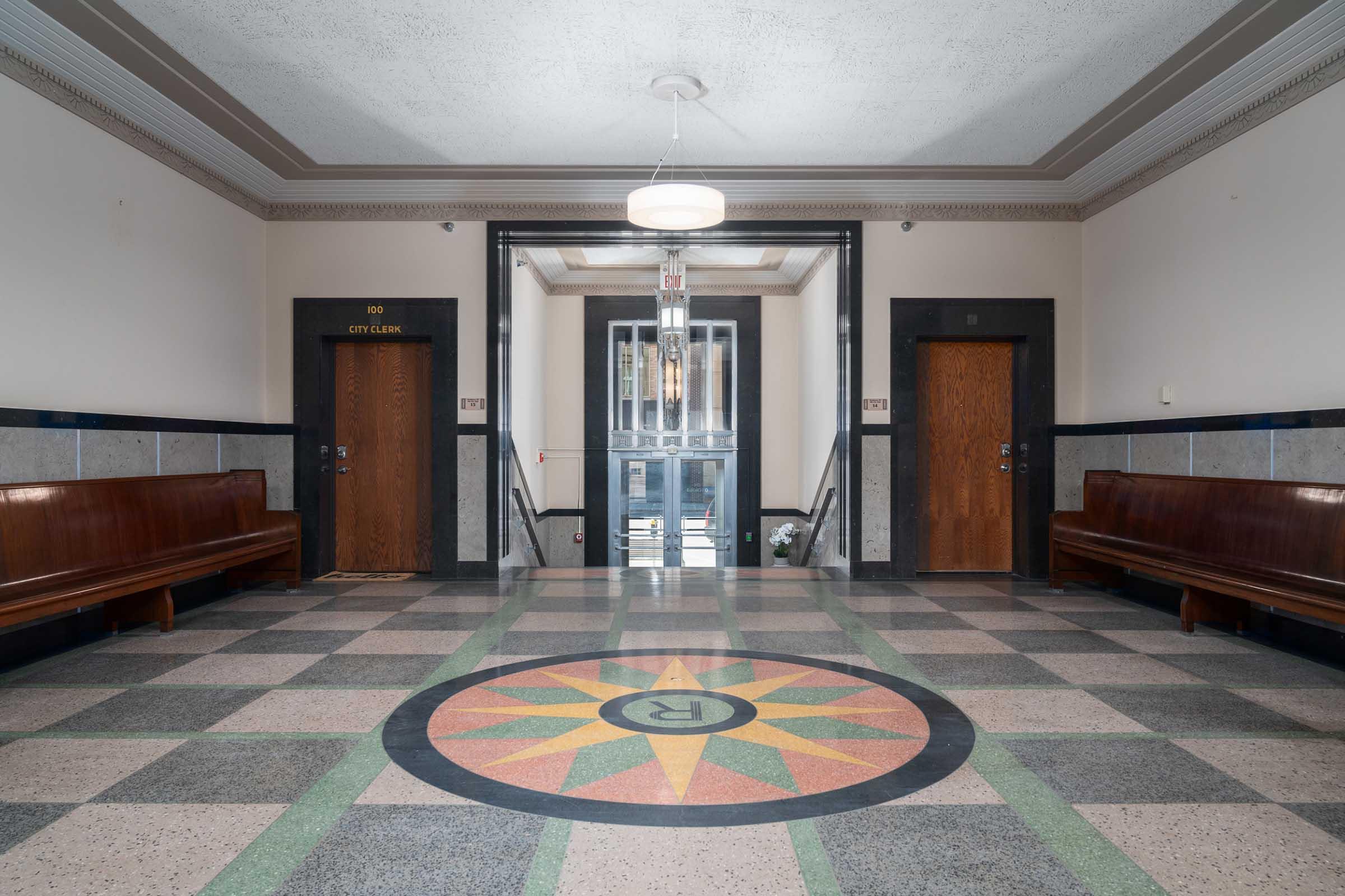 Interior view of a lobby featuring a circular compass rose design on the floor. Two wooden benches are positioned along the sides, with doors labeled "City Clerk" visible in the background. Natural light filters in from a ceiling fixture, highlighting the architectural details of the space.
