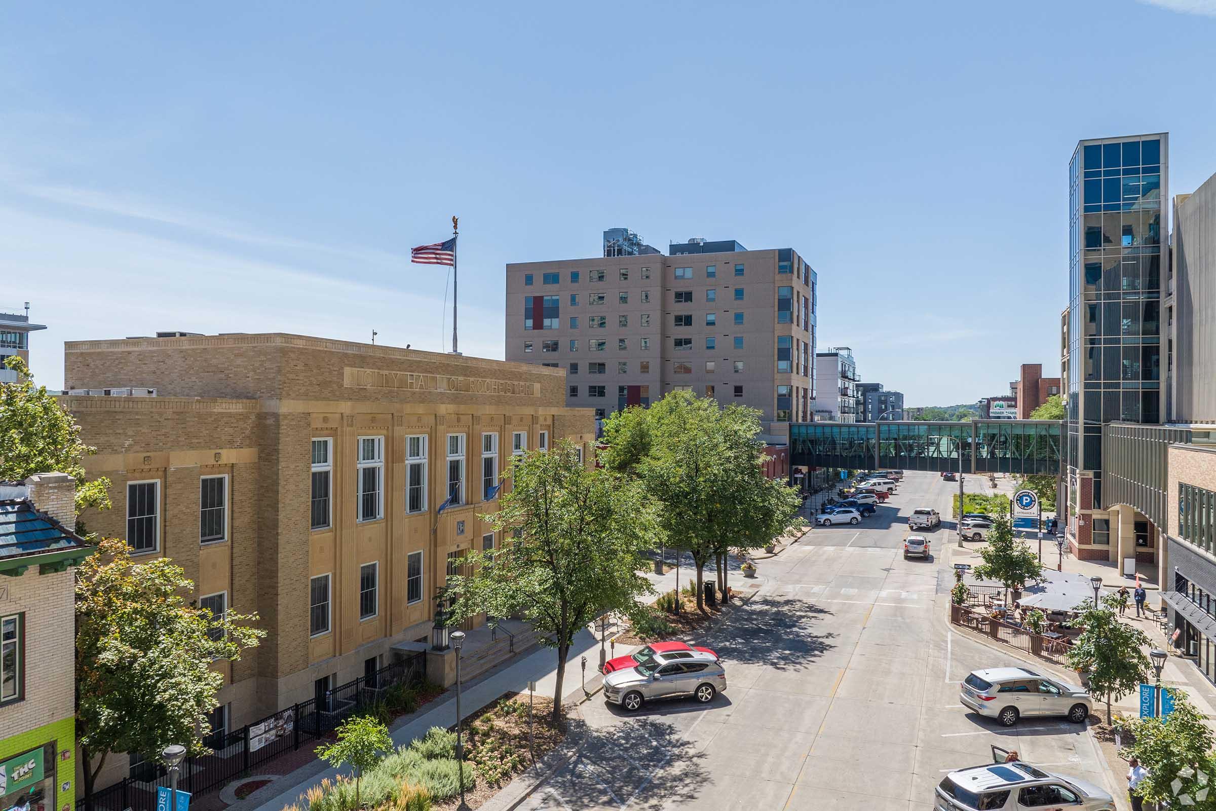 A clear view of a downtown city street lined with trees and parked cars. A historical brick building with an American flag is visible on the left, while modern buildings and a pedestrian bridge are seen in the background. The scene reflects a blend of contemporary and classic architecture under a bright blue sky.