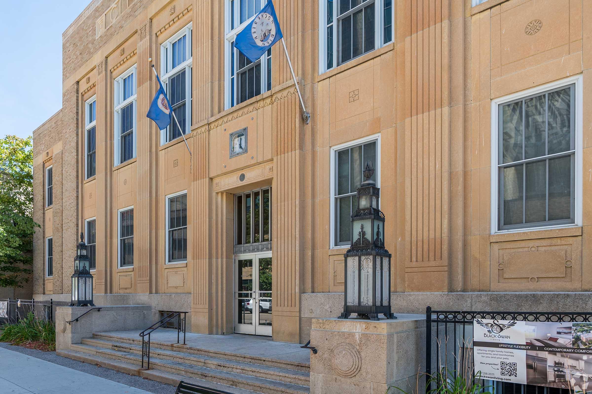Facade of a stone building with large windows and a central entrance. The entrance features steps leading up to double doors and is flanked by decorative lanterns. Two flags are flying above the entrance, and there is a sign on the side displaying information about nearby apartments.