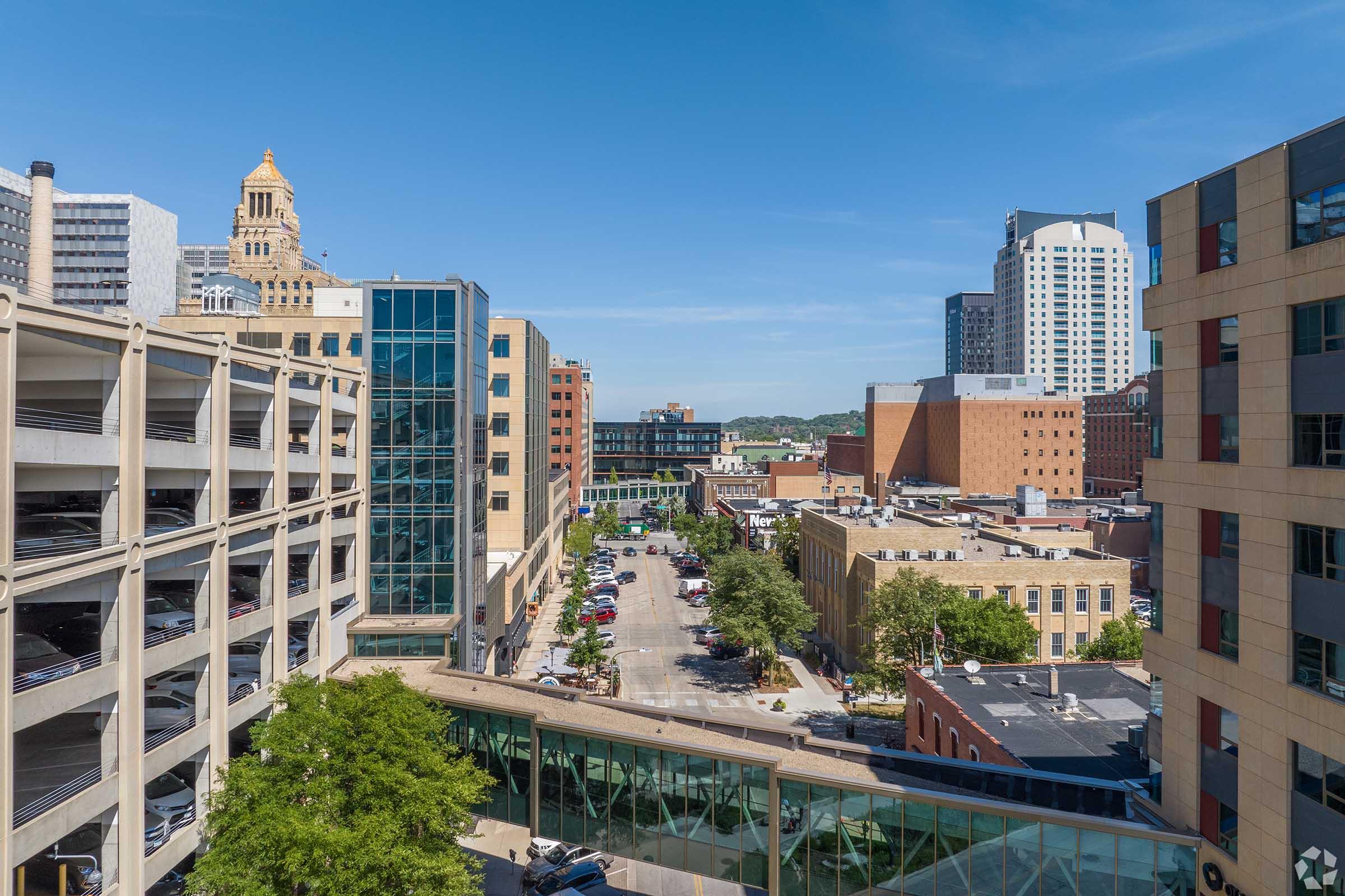 Aerial view of a downtown cityscape featuring a mix of modern and historical buildings, a parking structure on the left, and tree-lined streets filled with parked cars. The skyline showcases high-rises and a clear blue sky, contributing to a vibrant urban atmosphere.