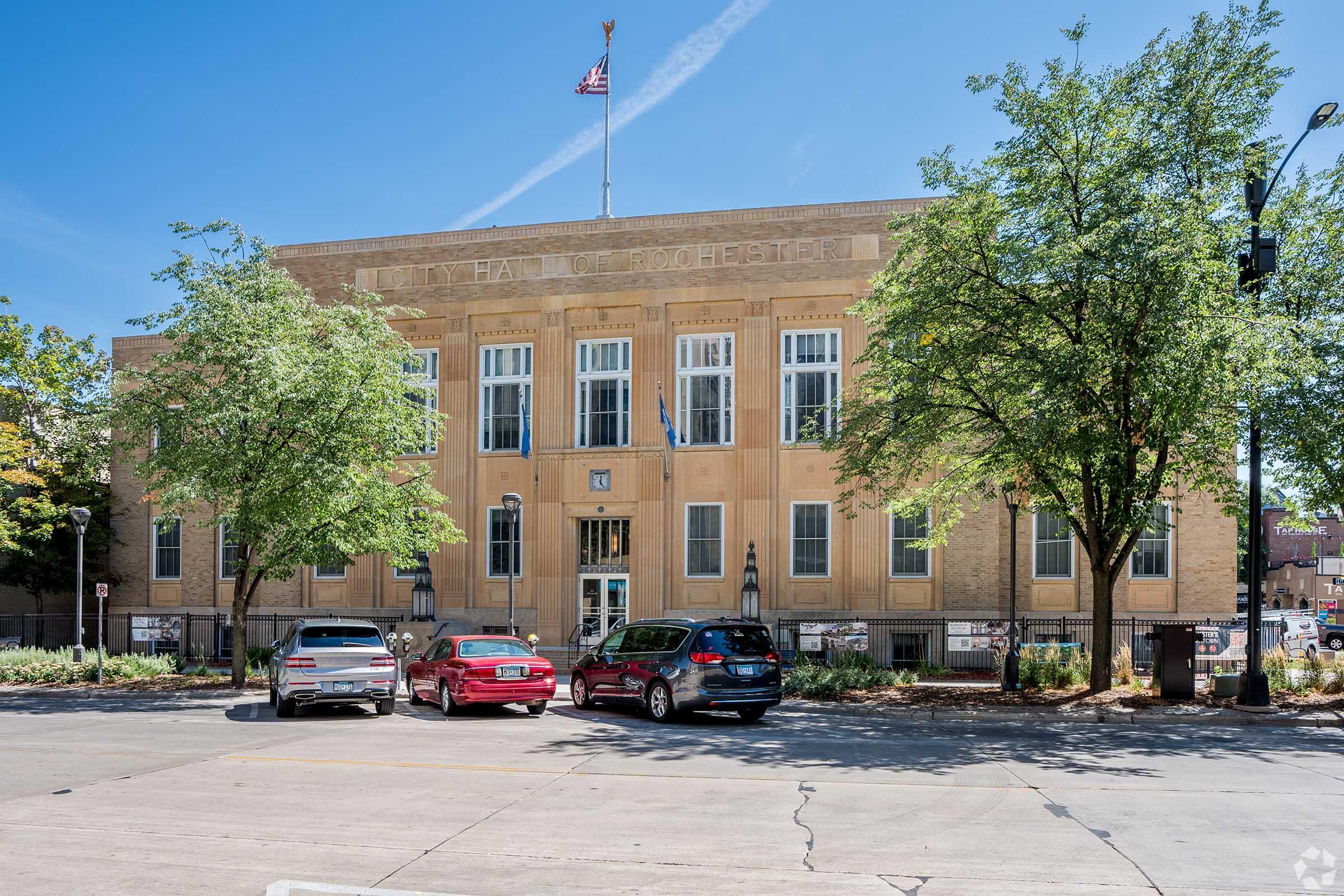 A large, historic building with a stone facade and multiple windows, featuring an American flag on a flagpole. In the foreground, several cars are parked in front of the building, and trees provide greenery around the area. Clear blue sky is visible above.