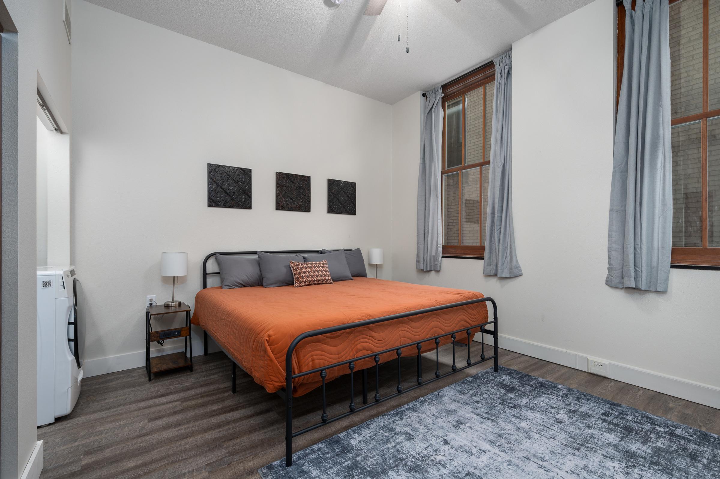 A cozy bedroom featuring a neatly made orange bed with gray bedding. Above the bed, there are three decorative wall hangings. The room has two large windows with gray curtains, and a bedside table with a lamp on one side. A washing machine is visible near the doorway, and the flooring is wooden with a large blue rug.