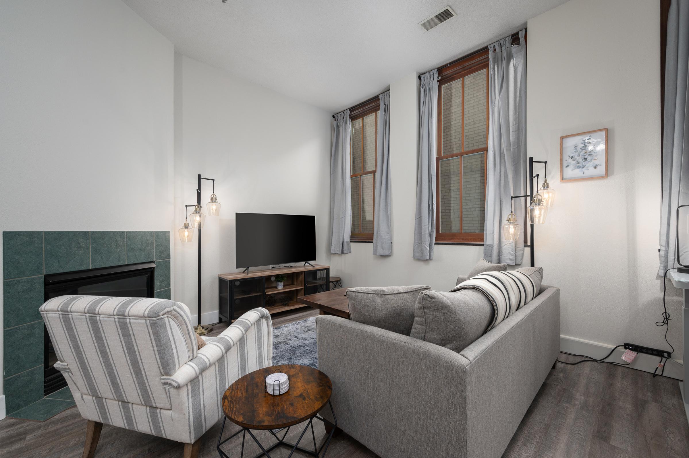 A cozy living room featuring a grey sofa and a striped armchair, a wooden coffee table, and a TV on a stand. The space has large windows with light grey curtains and a small table with a decorative item. There’s also a fireplace with teal tiles, creating a warm ambiance.
