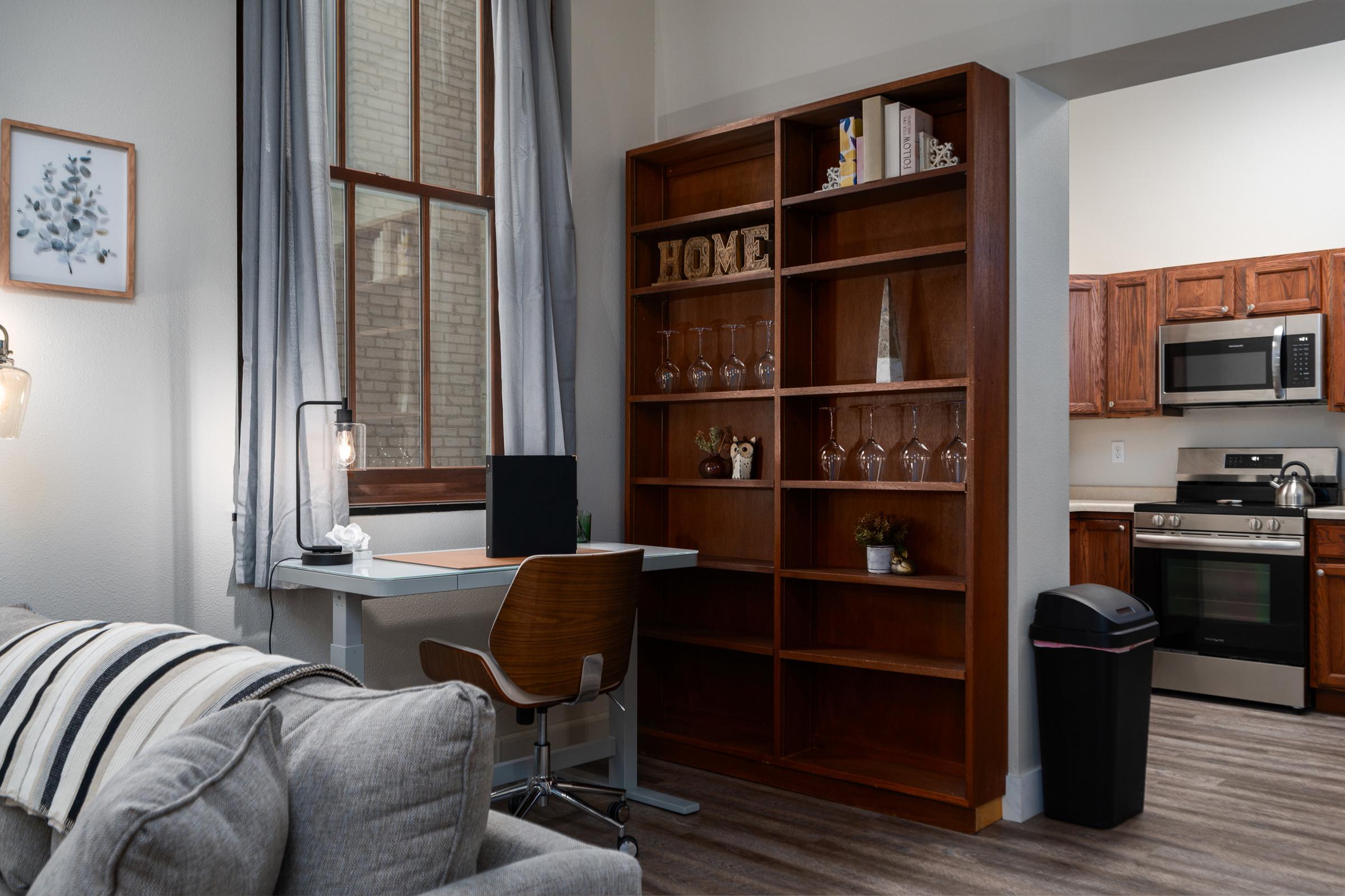 Cozy living space featuring a wooden bookshelf filled with glassware and decor, a modern desk with a laptop, and a comfortable sofa. The kitchen is visible in the background with stainless steel appliances, and large windows allow natural light to brighten the room.