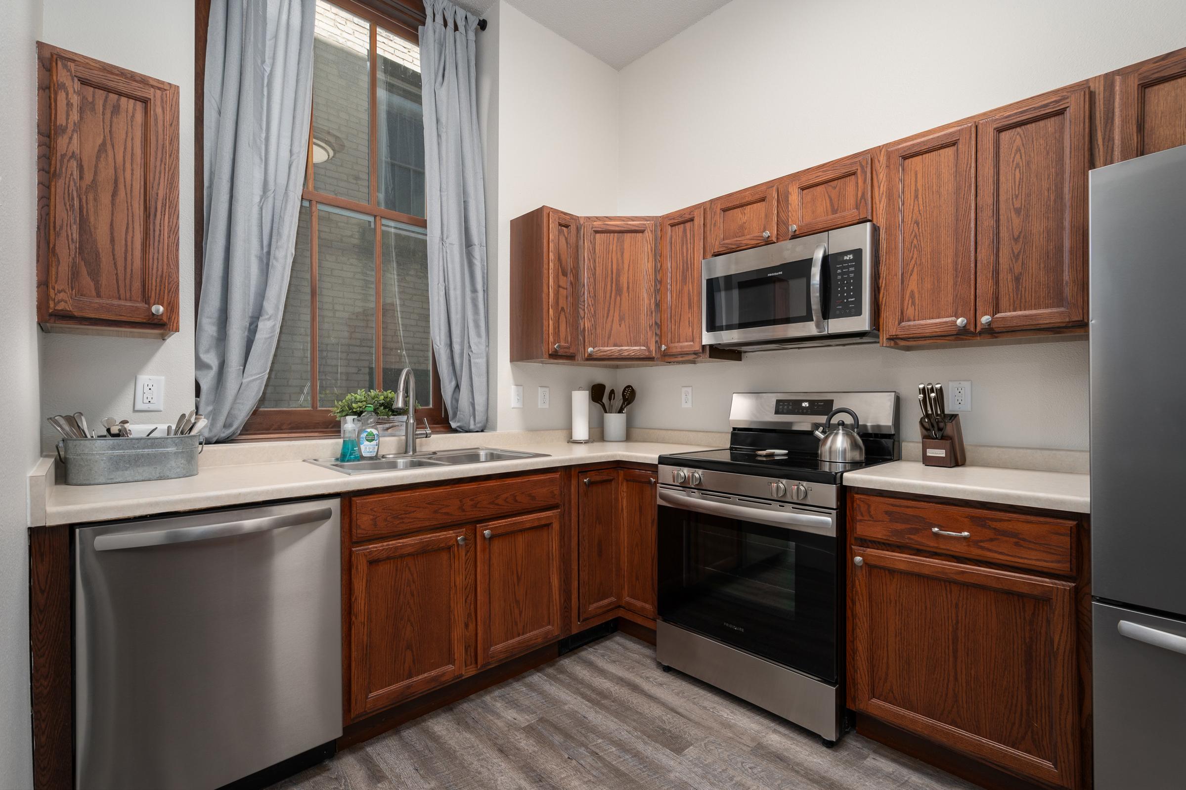 Modern kitchen featuring wooden cabinets, stainless steel appliances including a refrigerator and oven, a stainless steel sink, and large windows with curtains. The countertop is beige, and the flooring is gray. Kitchen utensils and a potted plant are visible on the counter.