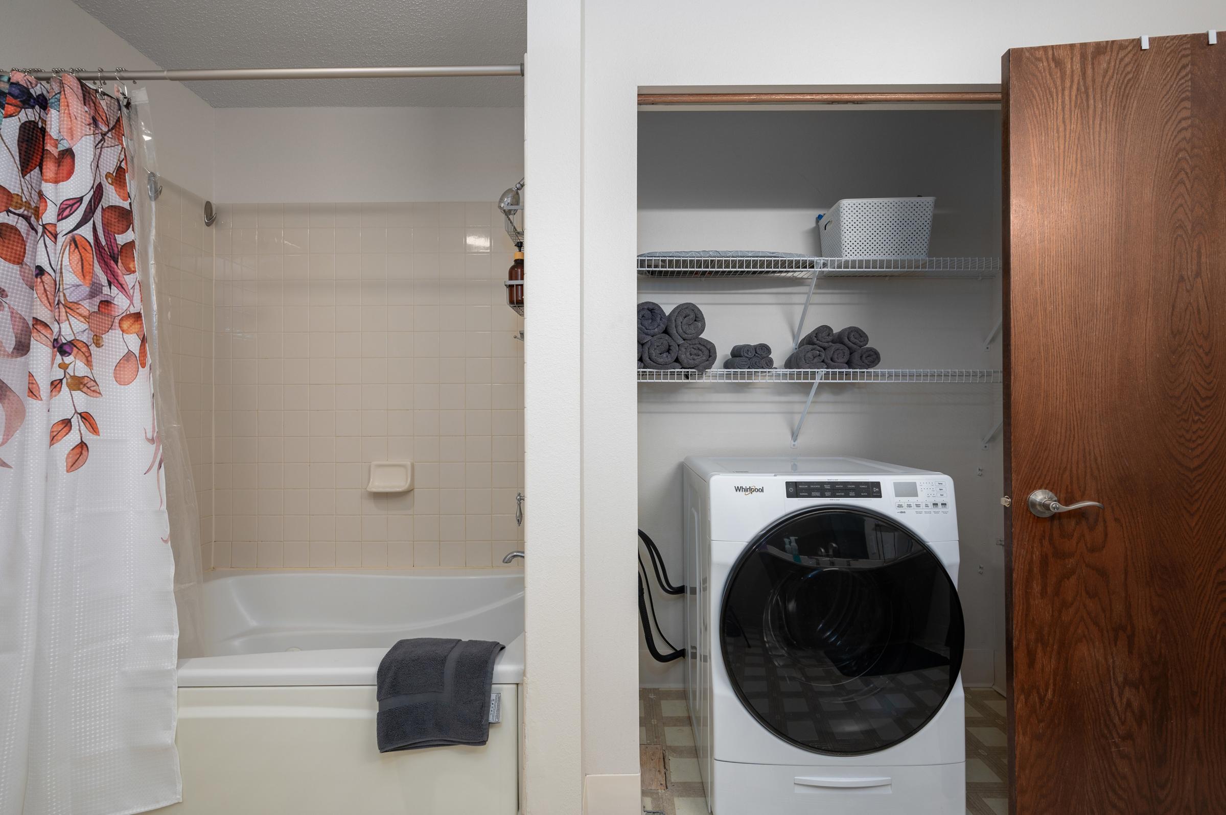 A bathroom with a bathtub on the left and a laundry area on the right, featuring a stacked washer and dryer. The bathroom has a floral shower curtain and a gray towel draped over the tub. The laundry area is organized with shelves holding rolled towels and a basket. The overall aesthetic is clean and minimalistic.