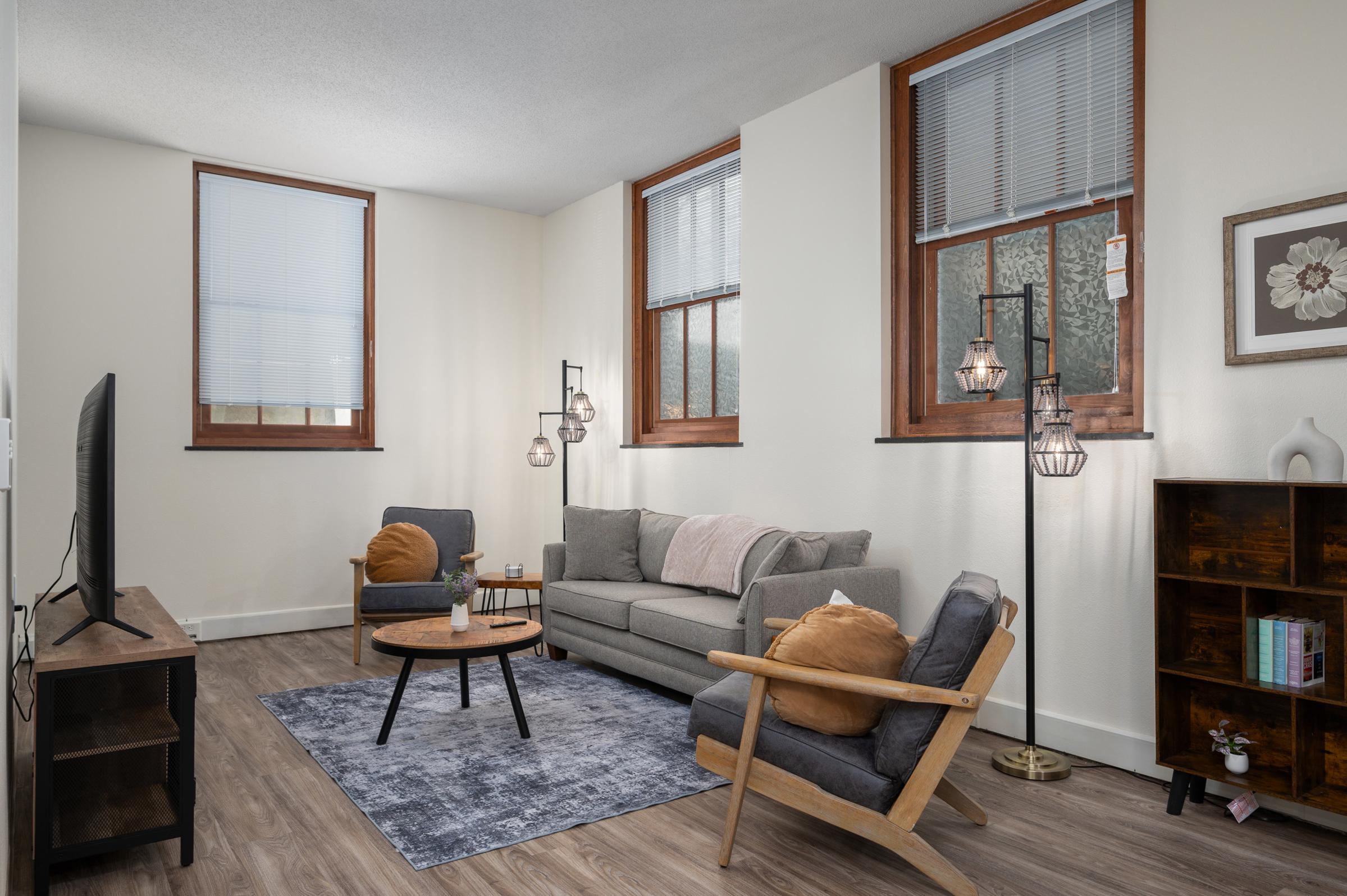 A cozy living room featuring a grey sofa, two armchairs, and a round coffee table. Natural light streams through two windows with blinds. A modern floor lamp and a bookshelf add a stylish touch. A decorative painting is visible on the wall, complementing the warm tones of the furnishings.