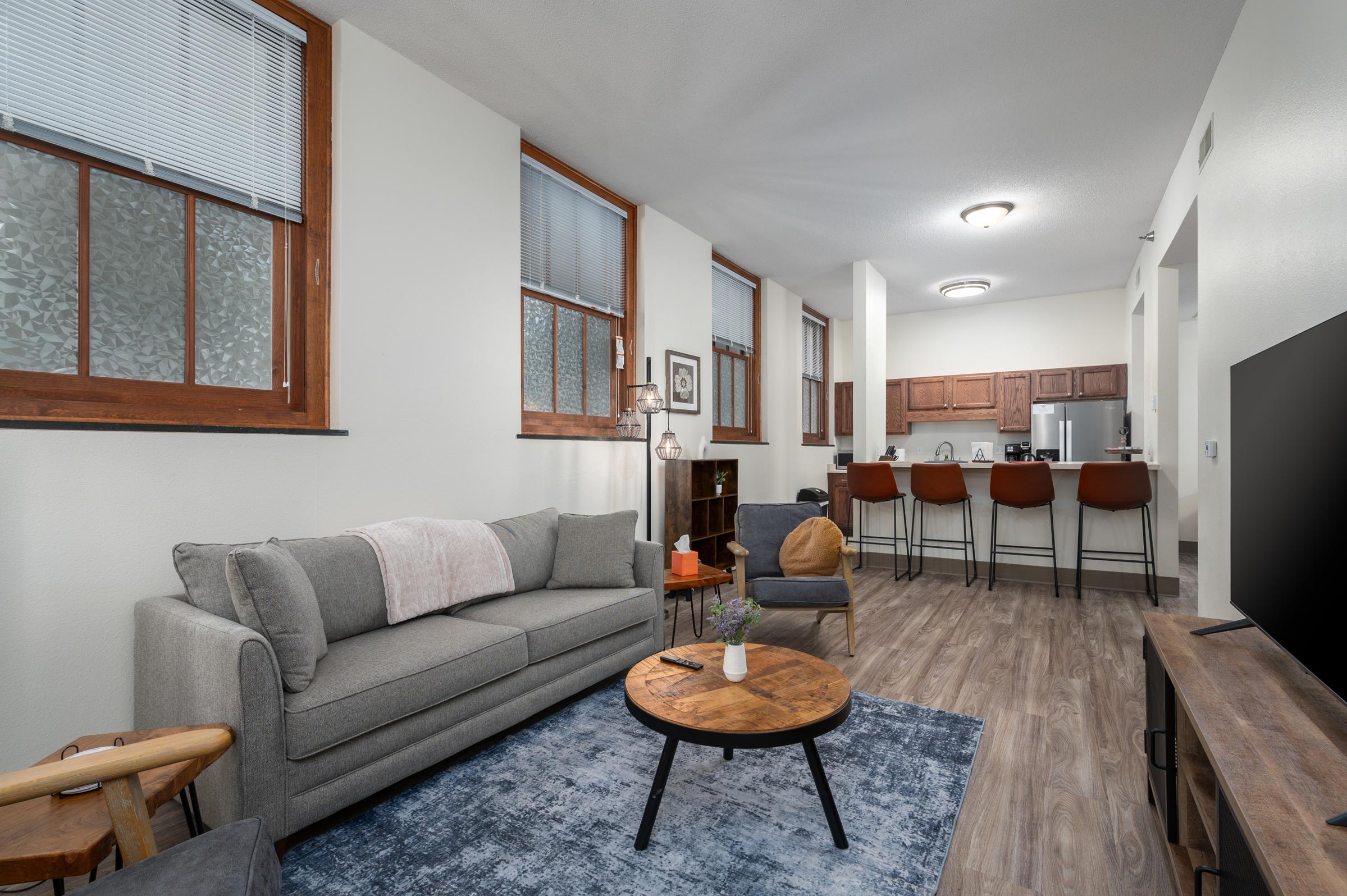 A cozy living room featuring a gray sofa with a throw pillow, a round wooden coffee table, and a flat-screen TV on a wooden stand. The space has large windows with natural light, modern kitchen bar stools visible in the background, and a warm, inviting atmosphere with neutral colors and decorative accents.