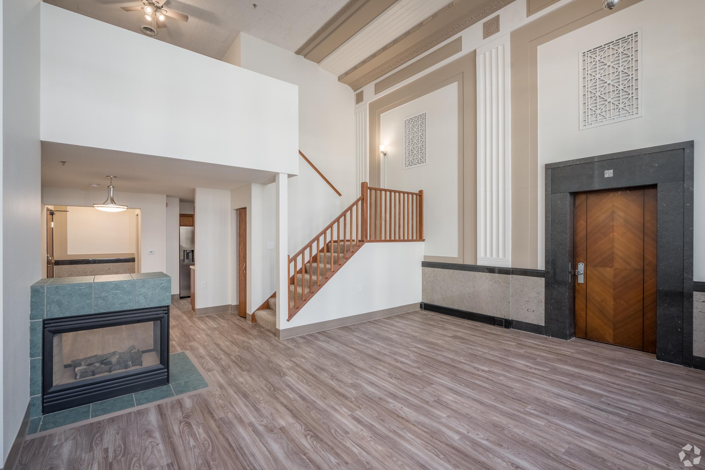 Interior view of a modern, spacious room featuring hardwood floors, a decorative fireplace, and a staircase. The walls have elegant detailing, and there is a well-lit area with a ceiling fan. Large windows provide natural light, creating an inviting atmosphere. The space is designed for comfort and style.