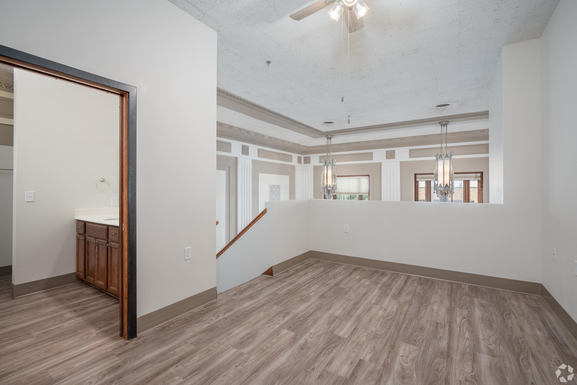 A spacious interior room featuring light-colored walls, decorative ceiling, and modern lighting fixtures. The floor has a wood-like appearance, and there is an open area leading to a staircase. An adjoining doorway leads to a bathroom with wooden cabinetry. Natural light brightens the space.
