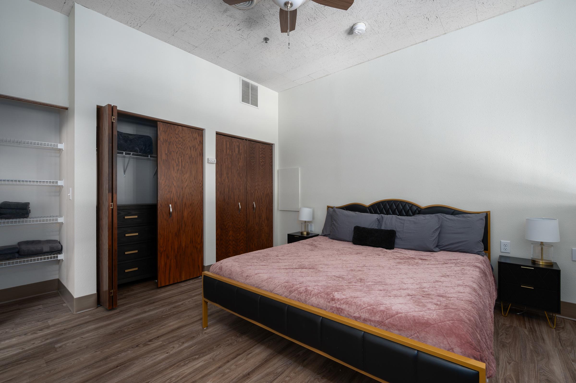 A cozy bedroom featuring a large bed with a pink blanket and gray pillows. There are two wooden closets with sliding doors, a black dresser with drawers, and two bedside lamps on either side of the bed. The walls are light-colored, and the flooring is a warm wood tone.