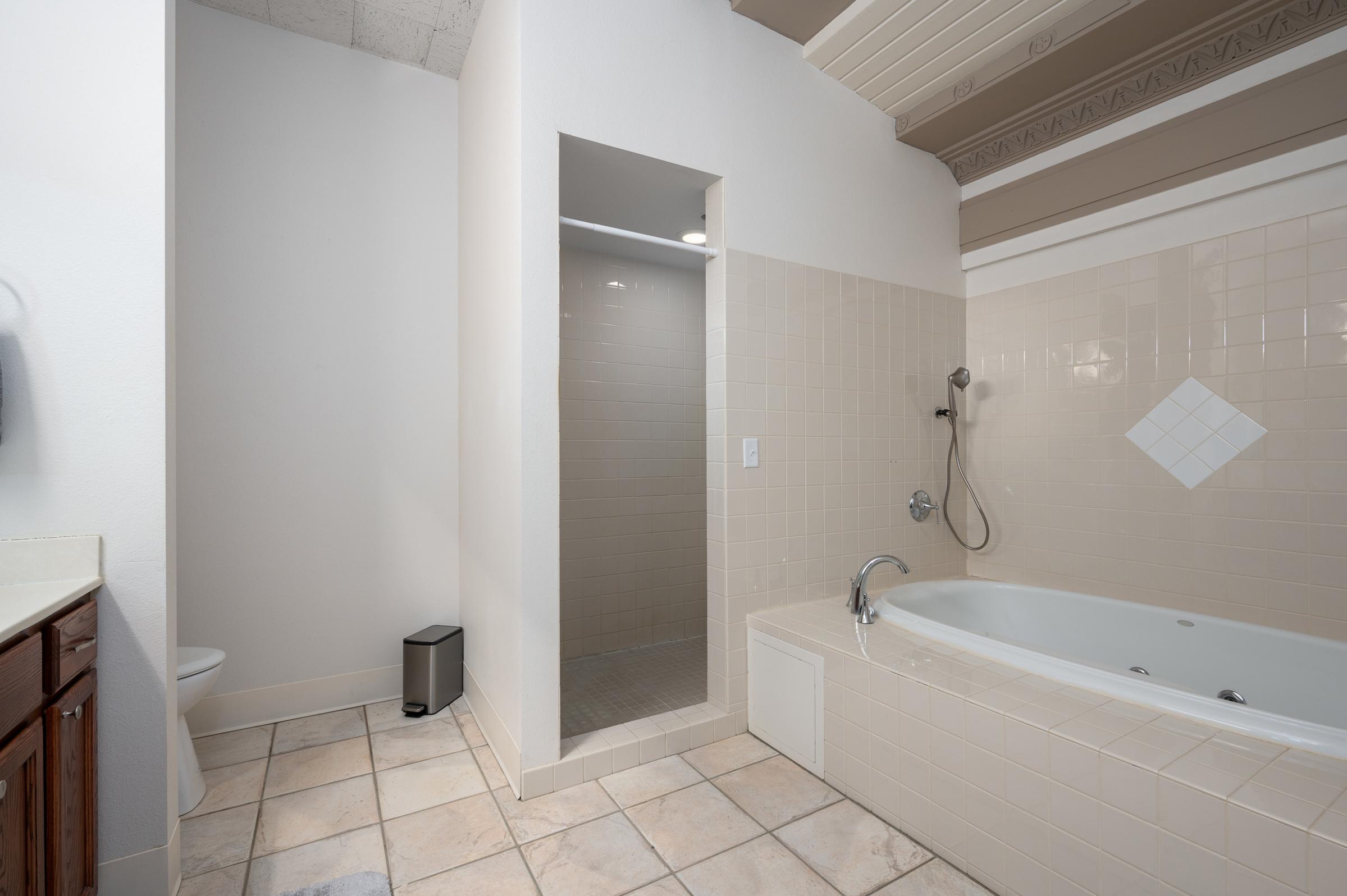 A spacious bathroom featuring a shower stall, a jetted tub, and a toilet. The walls are light-colored tiles, and the ceiling has decorative trim. The floor is tiled, and a small wastebasket is positioned near the shower area. Natural light fills the space, creating a bright atmosphere.