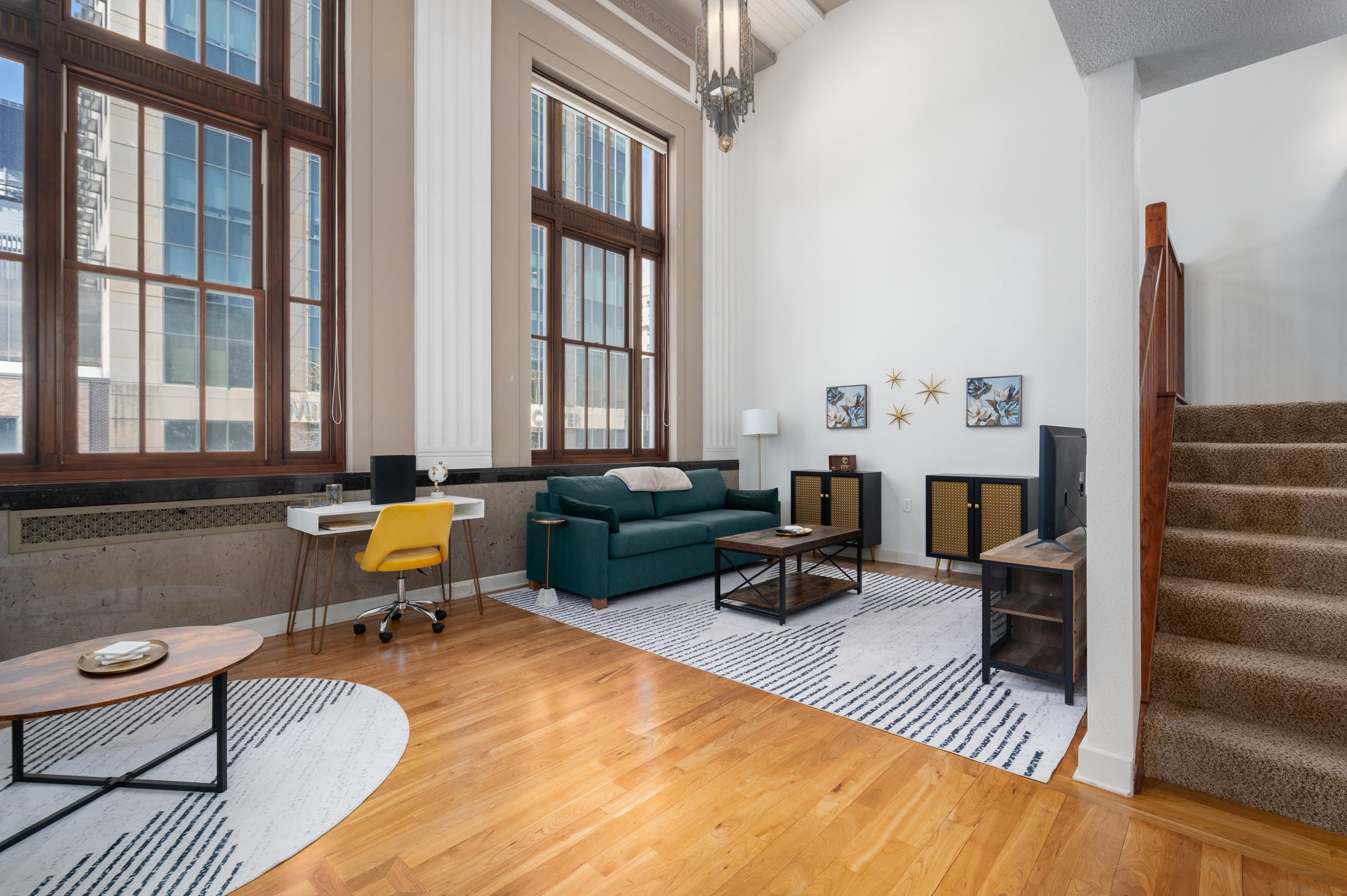 A bright and stylish living area featuring a green sofa, a yellow office chair, and modern decor. Large windows allow for natural light, highlighting wood and concrete elements. A staircase leads to an upper level, while a circular area rug adds softness to the wooden floor. Decorative wall art enhances the space.