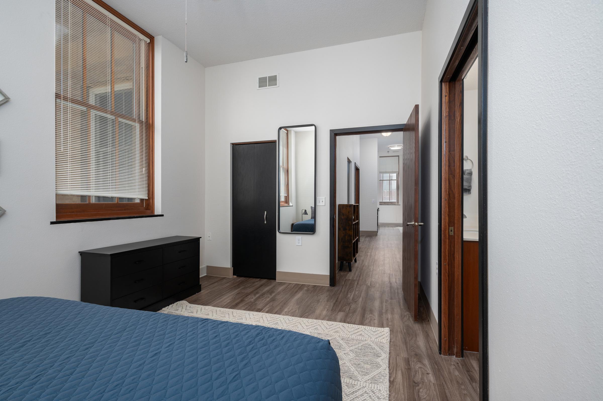 A modern bedroom featuring a dark blue quilted bedspread, a black dresser, and a mirror on the wall. Light-colored walls and wooden trim enhance the room's brightness. A doorway leads to a hallway, showing additional rooms with light flooring and neutral decor.