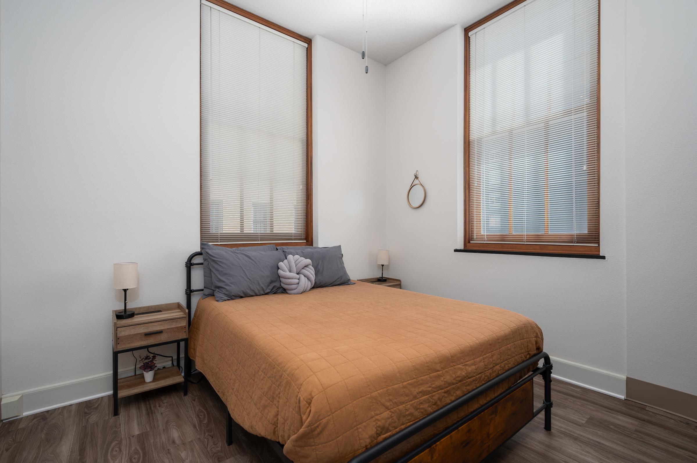 A cozy bedroom featuring a queen-size bed with an orange quilt, two bedside tables with lamps, and windows with blinds. The walls are painted white, and there’s a small mirror hanging on the wall. The floor is wooden, contributing to a warm, inviting atmosphere.