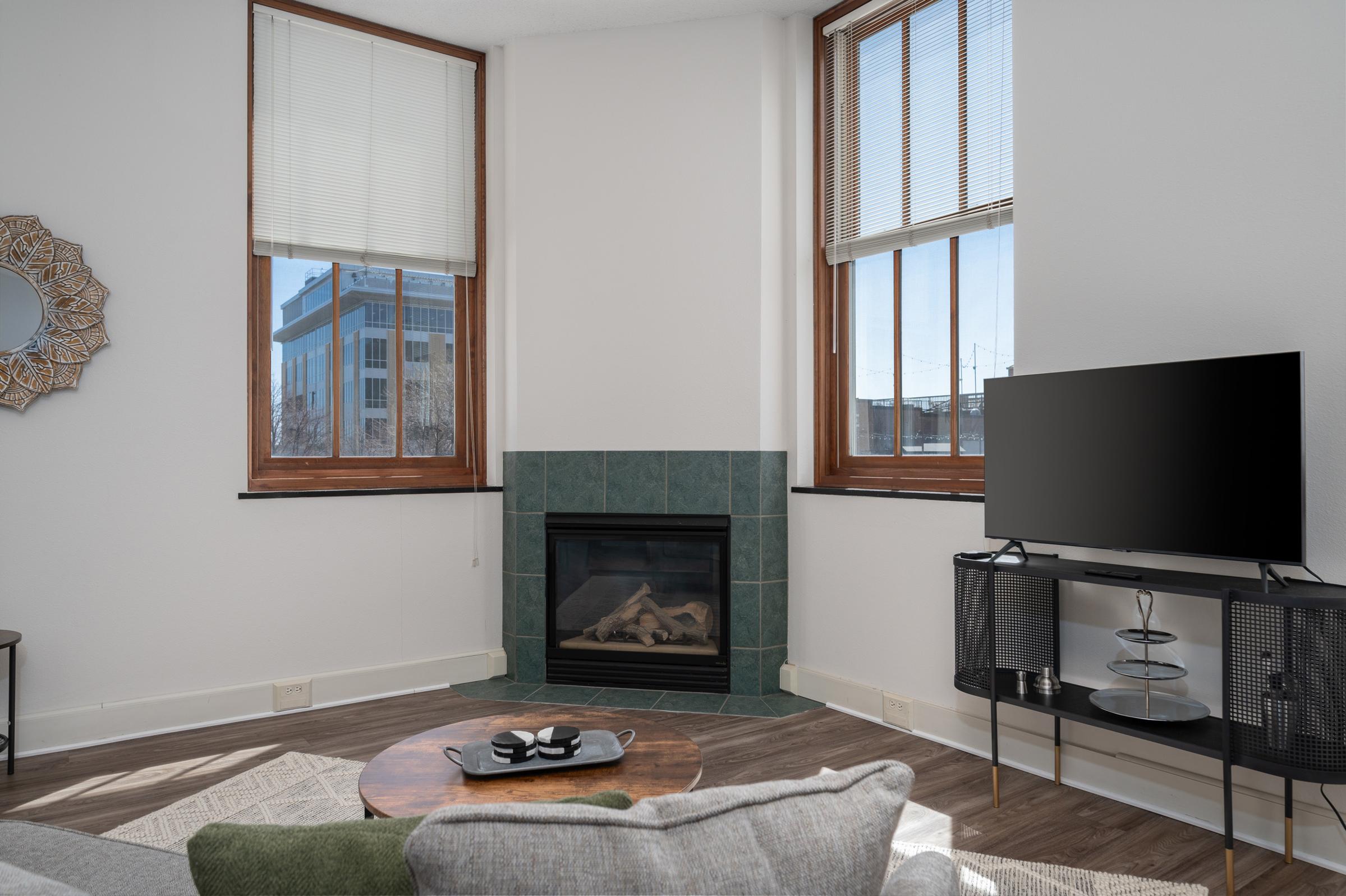 Bright living room with two large windows, a cozy fireplace with green tile, a circular wooden coffee table, a comfortable gray sofa with patterned cushions, and a TV on a black stand. Sunlight fills the space, highlighting the modern decor and inviting atmosphere.