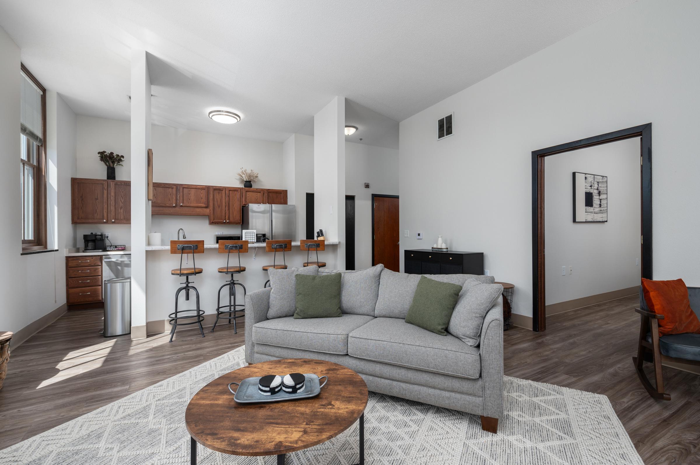 A modern living room and kitchen area featuring a gray sofa with decorative pillows, a round coffee table, and wooden bar stools at the kitchen counter. The space has large windows, wood-style flooring, and light-colored walls, creating a bright and inviting atmosphere.