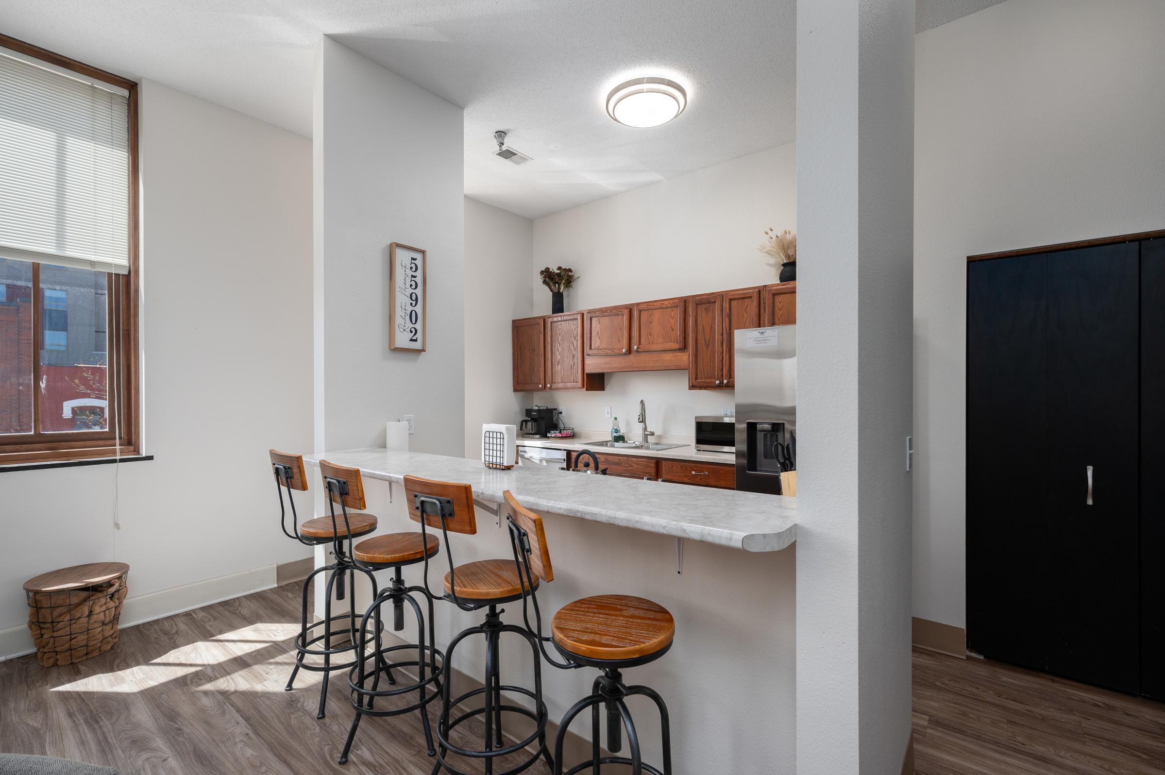 A modern kitchen with wooden cabinets and stainless steel appliances, featuring a white countertop with four barstools. Natural light streams in through a window, illuminating the space. A dark cabinet stands in the corner, adding contrast to the light decor.