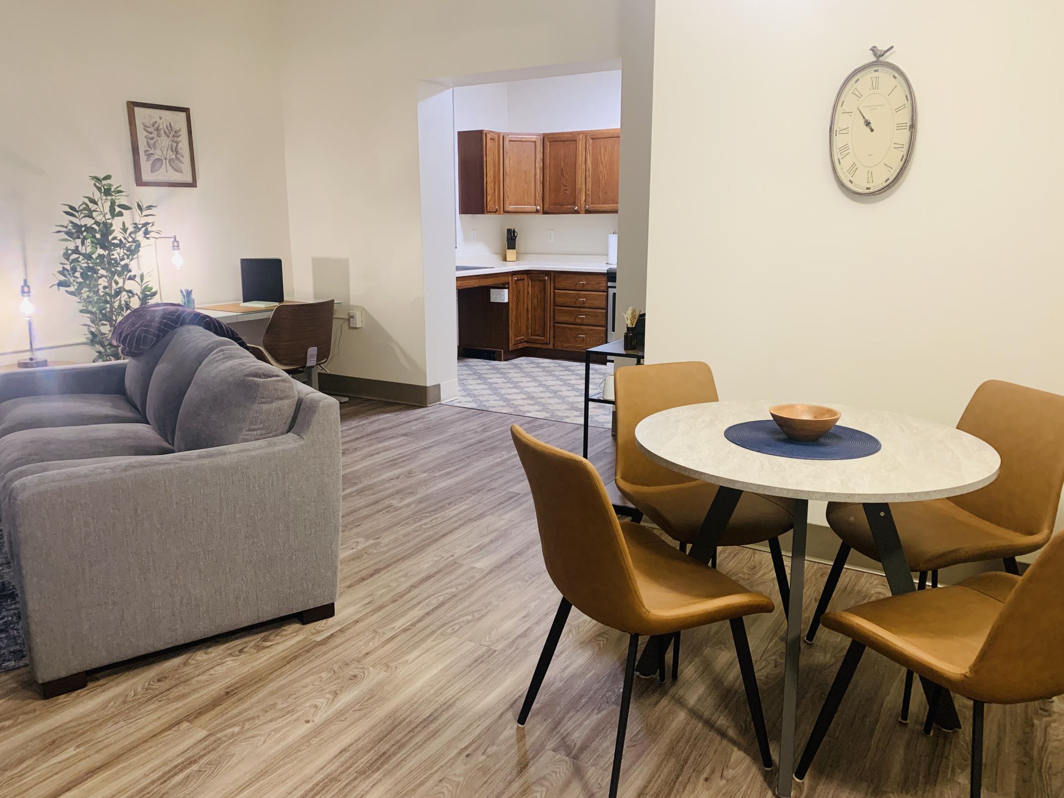 A cozy living area featuring a gray sofa and a round dining table with four brown chairs. A clock is mounted on the wall, and the space has light wood flooring. In the background, a kitchen with wooden cabinets is visible, adding to the homey atmosphere. Plants and lamps enhance the inviting ambiance.
