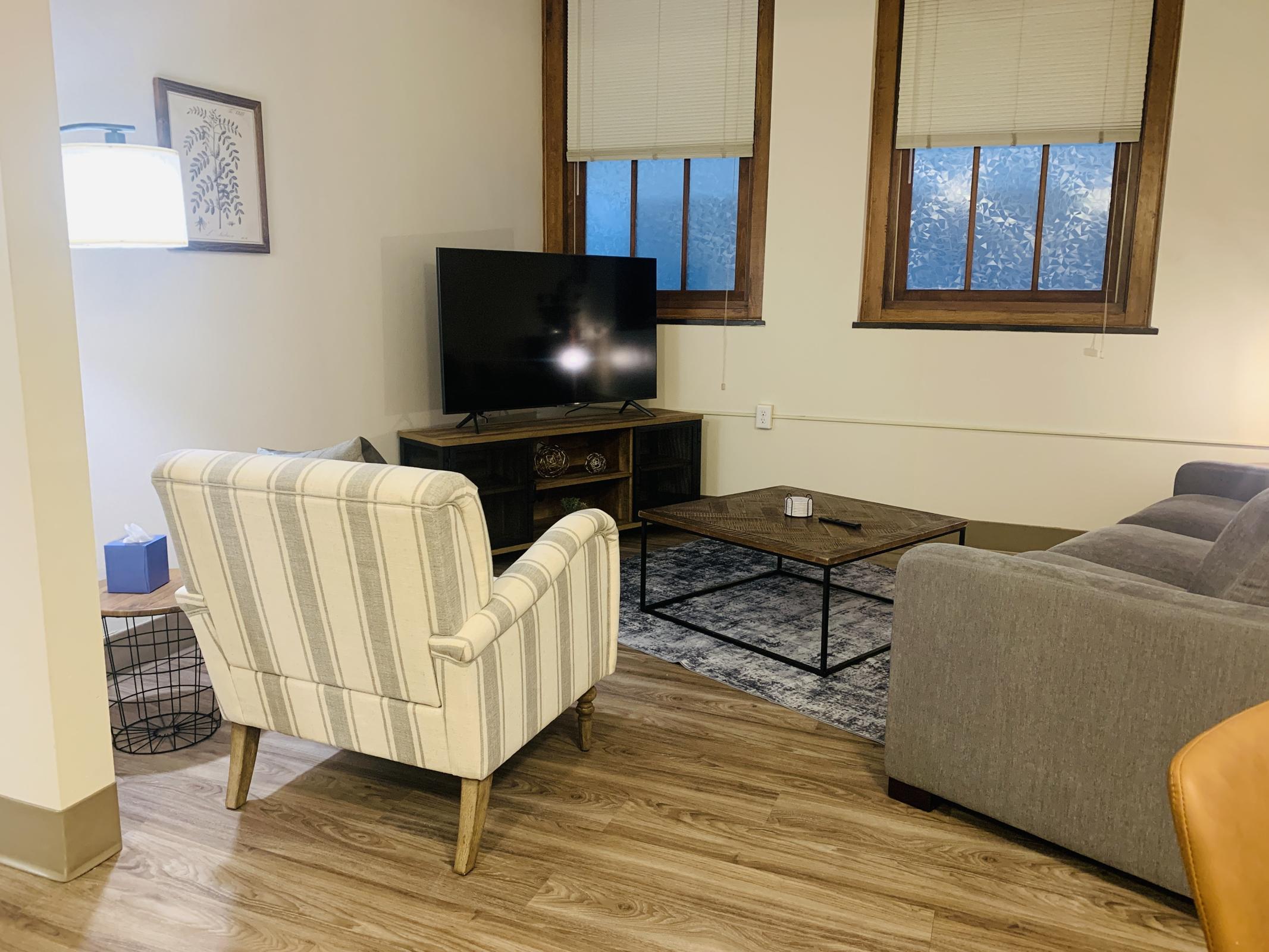 A cozy living room featuring a comfortable striped armchair, a gray sofa, a coffee table on a patterned rug, and a television on a wooden stand. Natural light comes through the windows, creating a warm atmosphere. A stylish lamp is positioned nearby, enhancing the inviting ambiance.