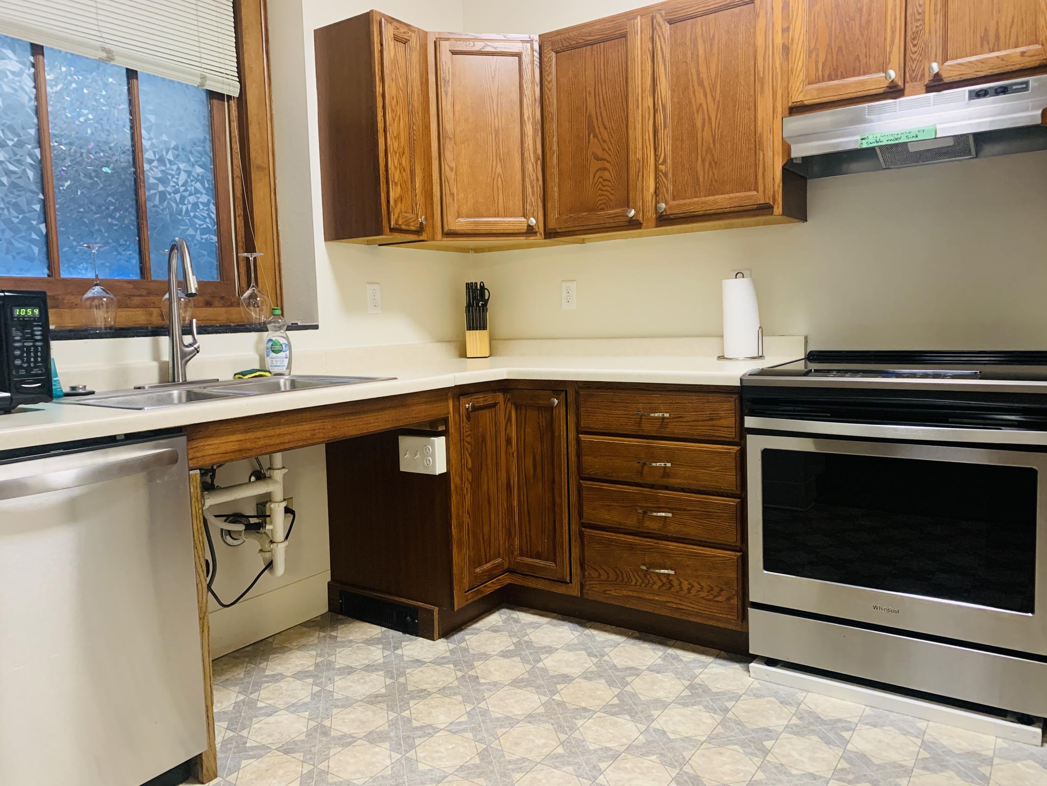 A modern kitchen with wooden cabinets, a stainless steel dishwasher, a sink with a faucet, and a black oven. The countertop is light-colored, and there's a window with natural light. The floor has a geometric pattern, and kitchen utensils are stored in a holder next to the sink.
