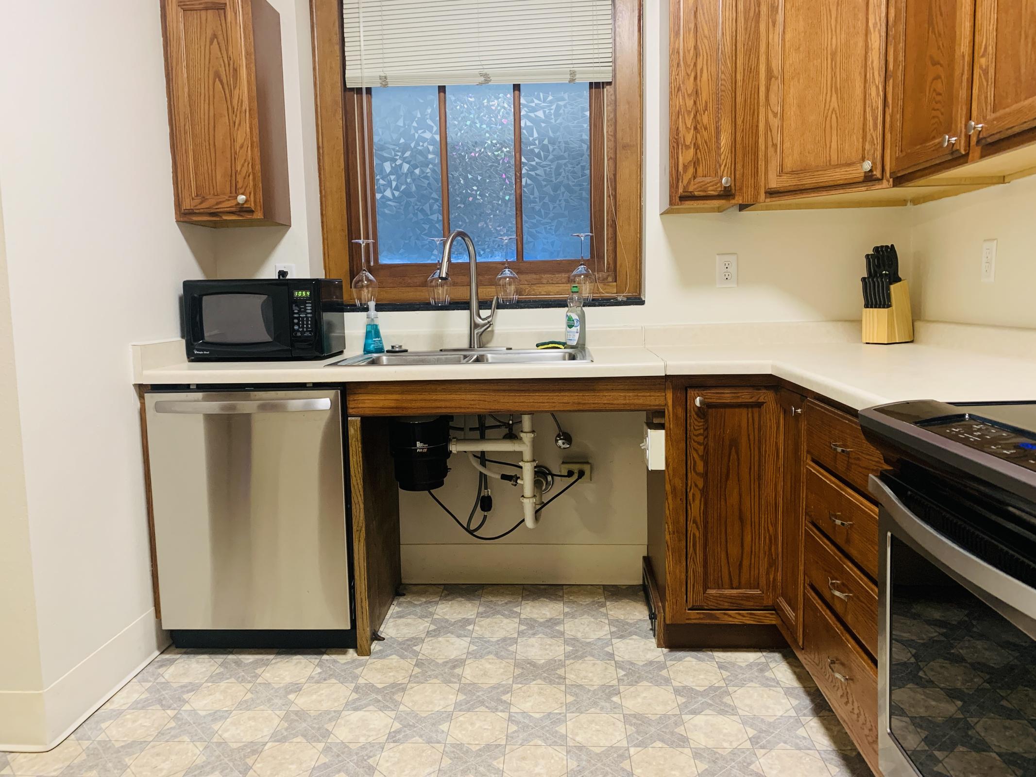 A modern kitchen featuring wooden cabinets, a stainless steel microwave, a silver dishwasher, and a black oven. The sink is situated under a window with frosted glass, and there are cleaning supplies on the countertop. The flooring has a geometric pattern, adding a stylish touch to the space.